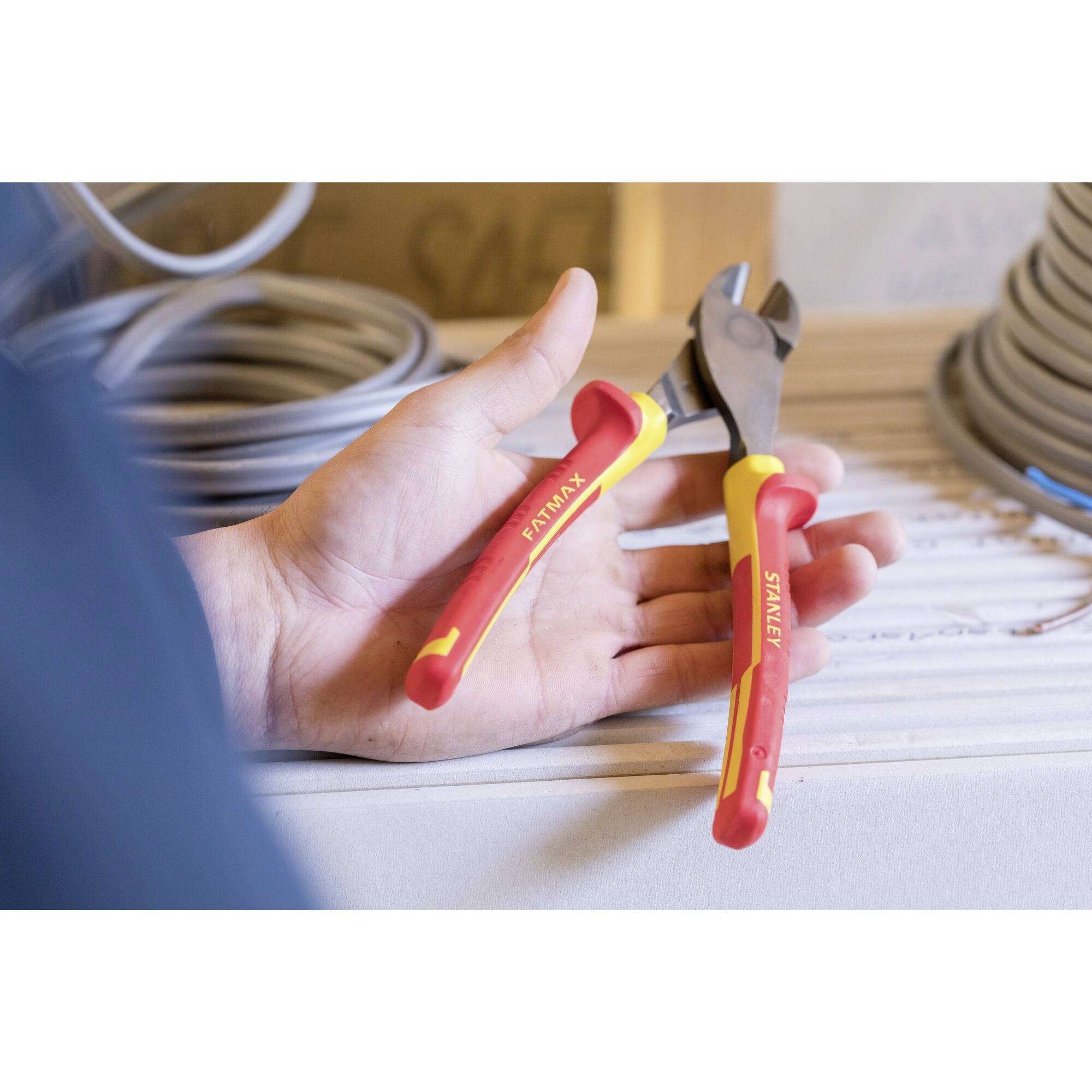 Une main tient une pince multifonction rouge avec une poignée jaune, tandis que des bobines de câbles sont visibles sur une table en arrière-plan.