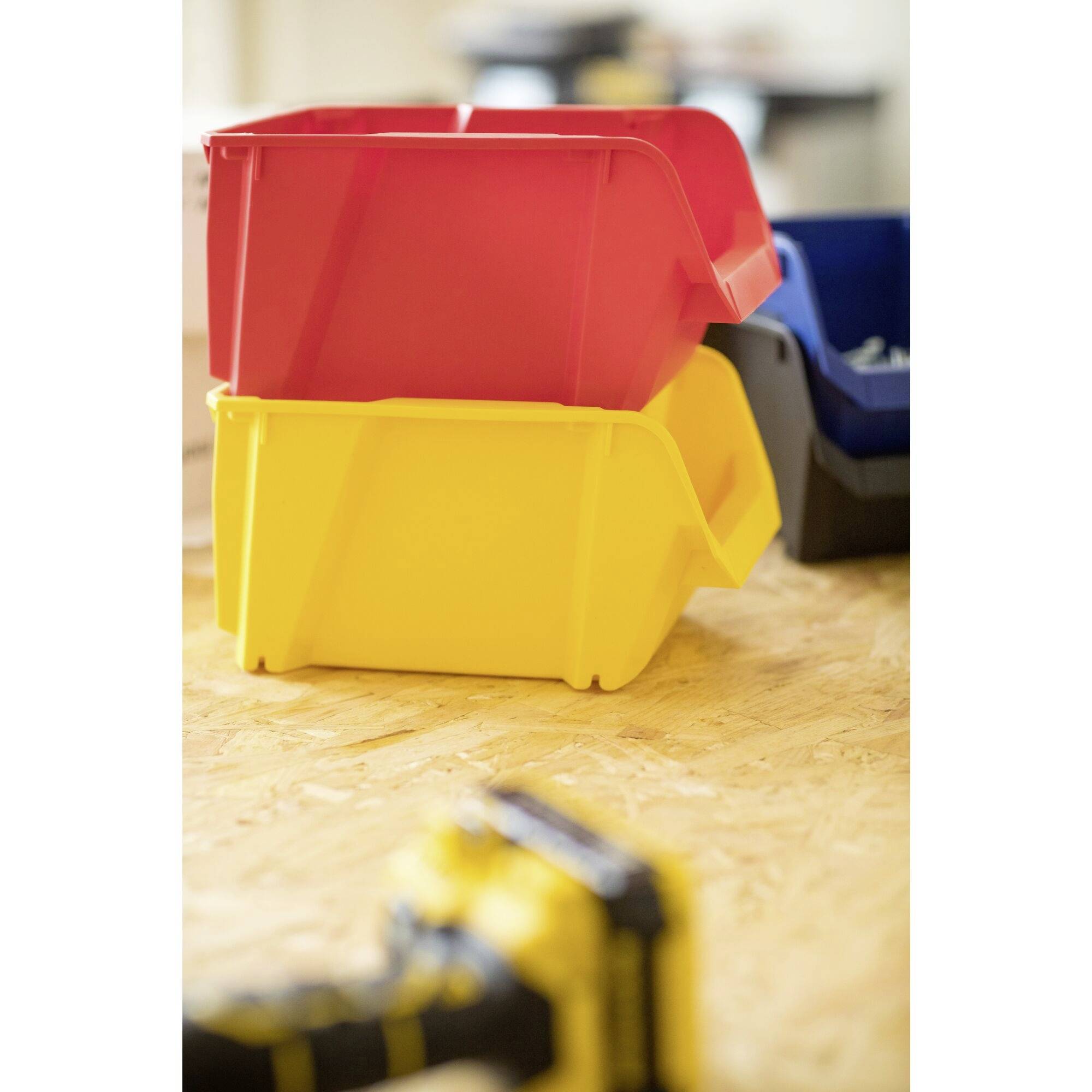 Trois boîtes de rangement empilées en rouge, jaune et bleu, sur une table en bois. La boîte jaune est au premier plan, cachant partiellement un outil flou.