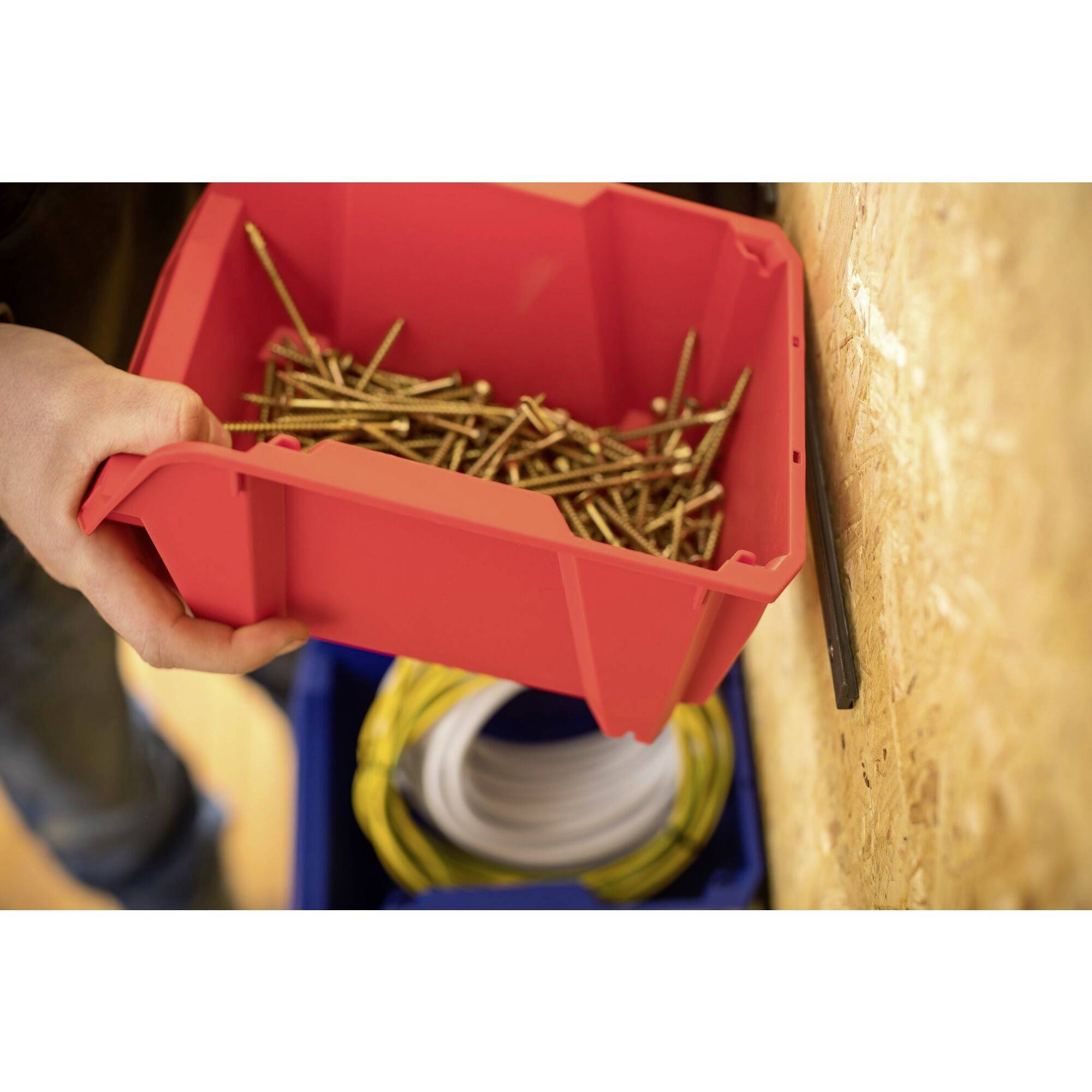 Une personne tient un récipient rouge rempli de longues vis dorées, devant un boîtier bleu avec un rouleau de câble jaune en arrière-plan.