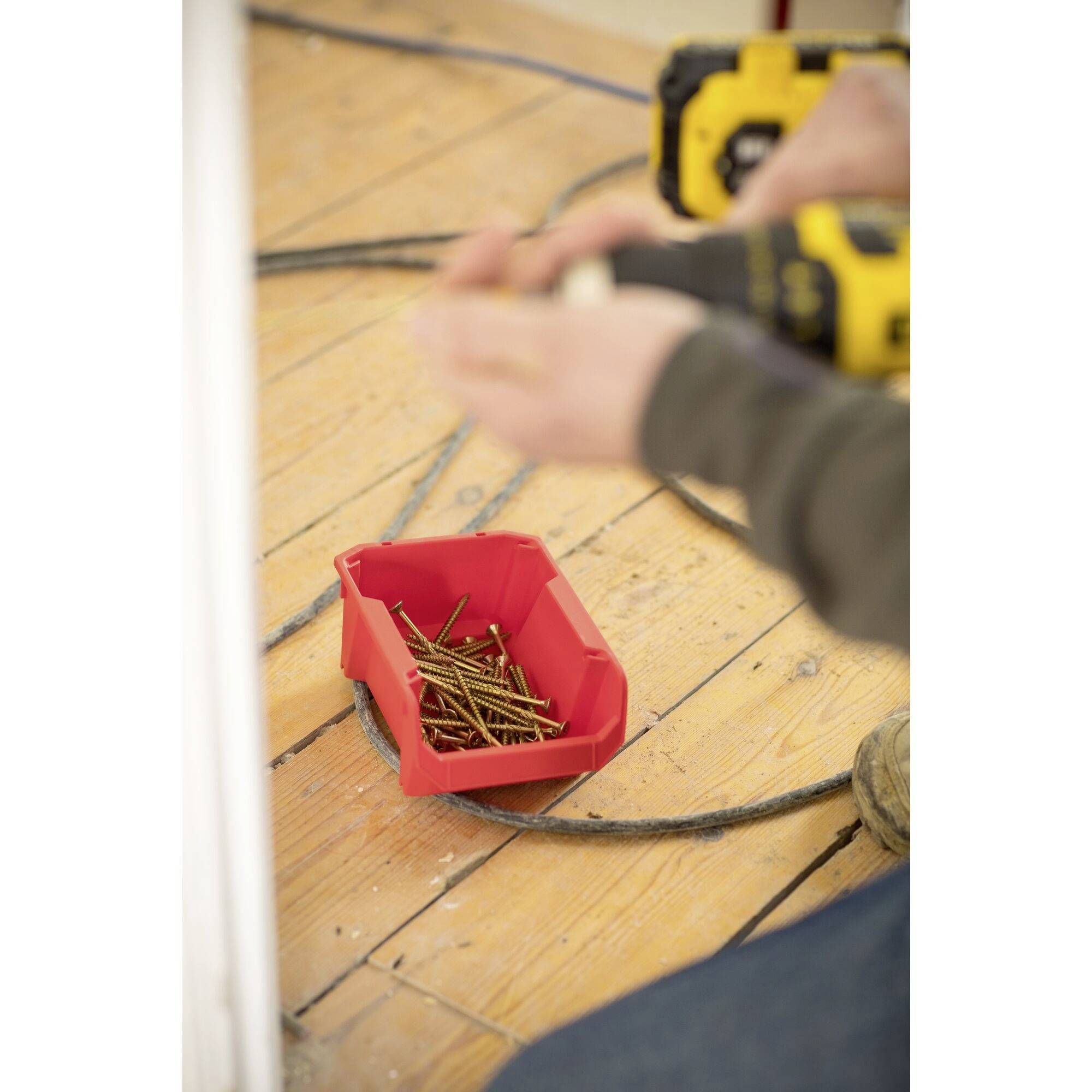 Une personne tient une perceuse au-dessus d'un plancher en bois. Au premier plan, se trouve un bol rouge contenant des vis dorées.