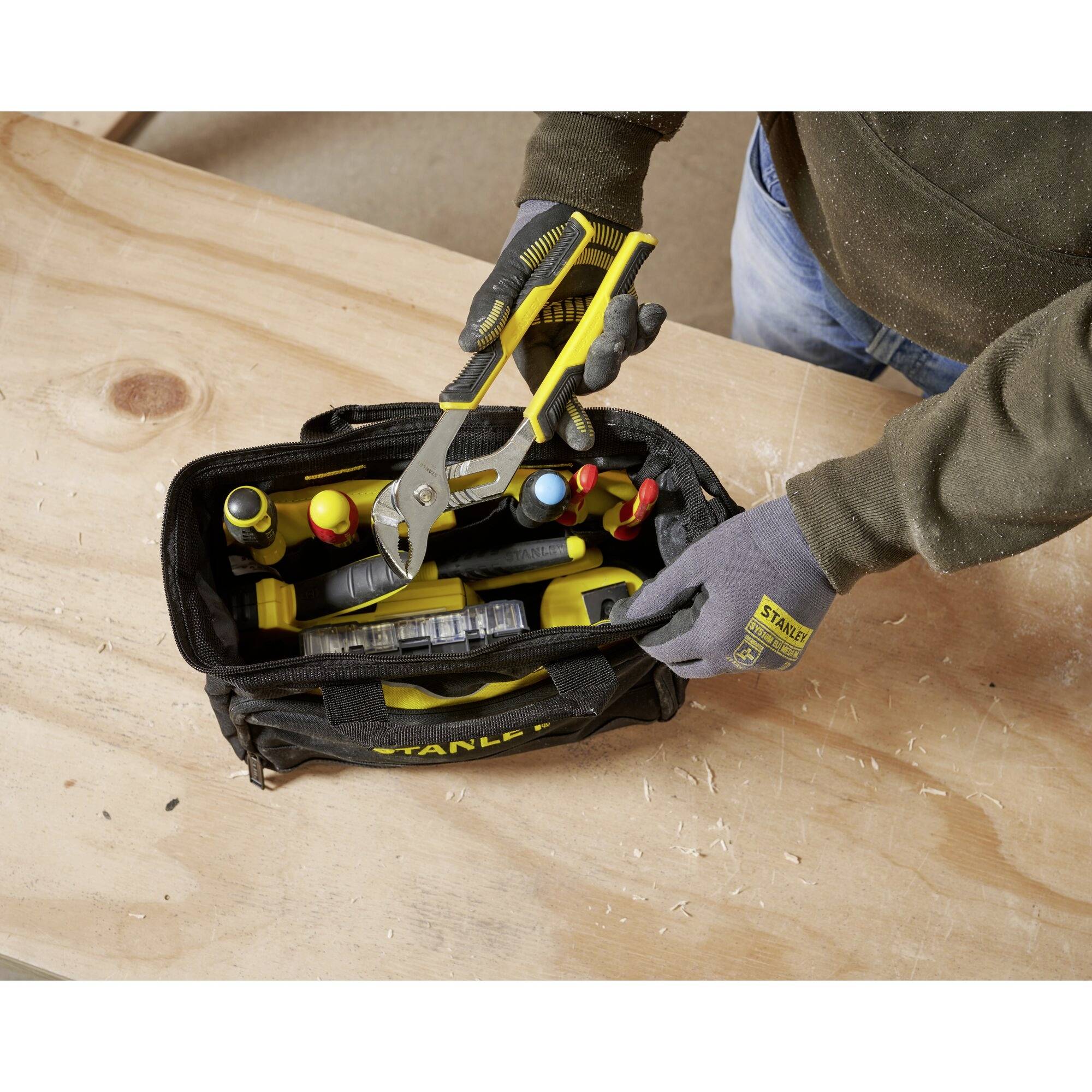 Une personne range une pince dans un sac à outils. Le sac contient divers outils. Il est posé sur une surface en bois.