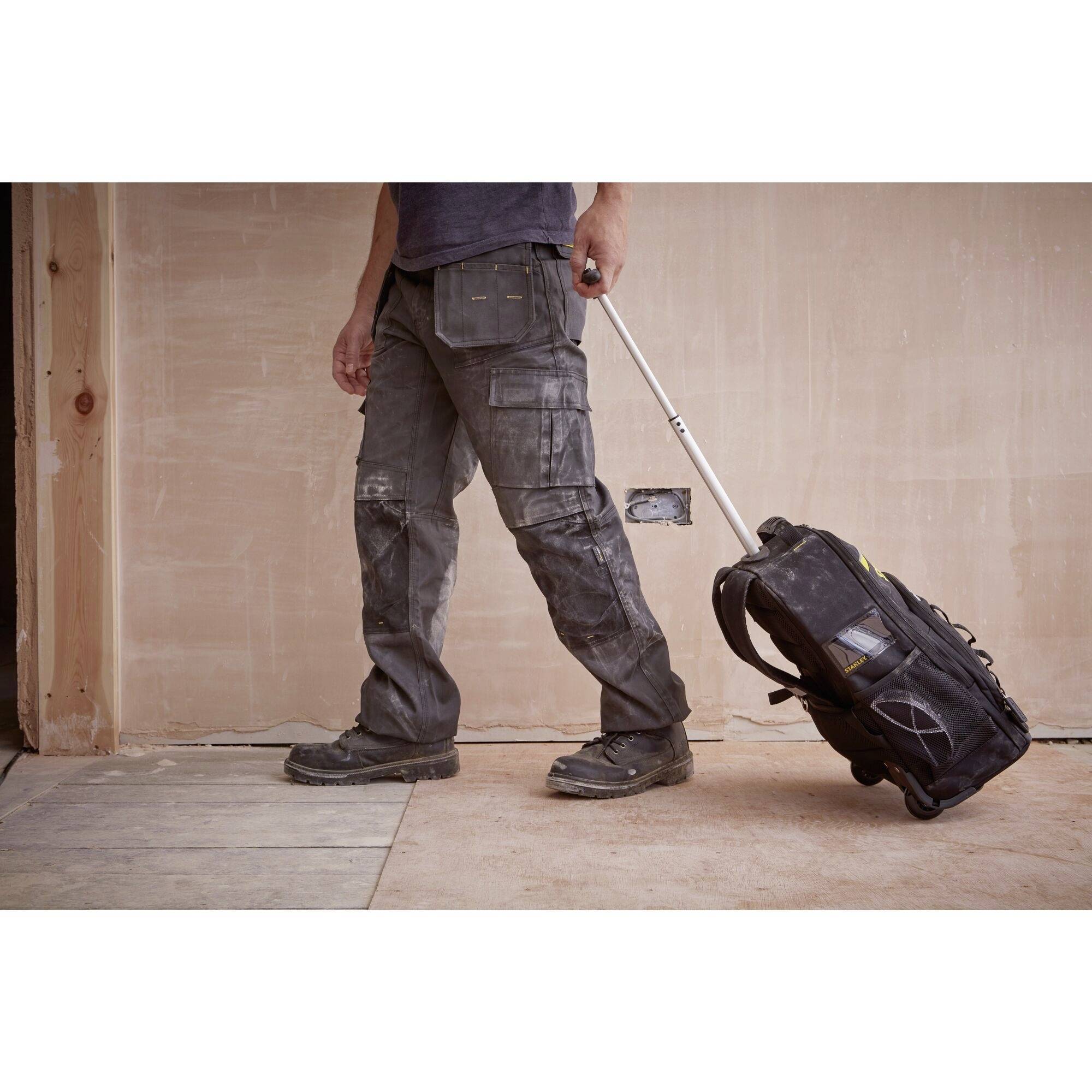Une personne tire un sac à dos à roulettes sur un plancher en bois de construction. La personne porte des vêtements de travail et est visible de la taille vers le bas.