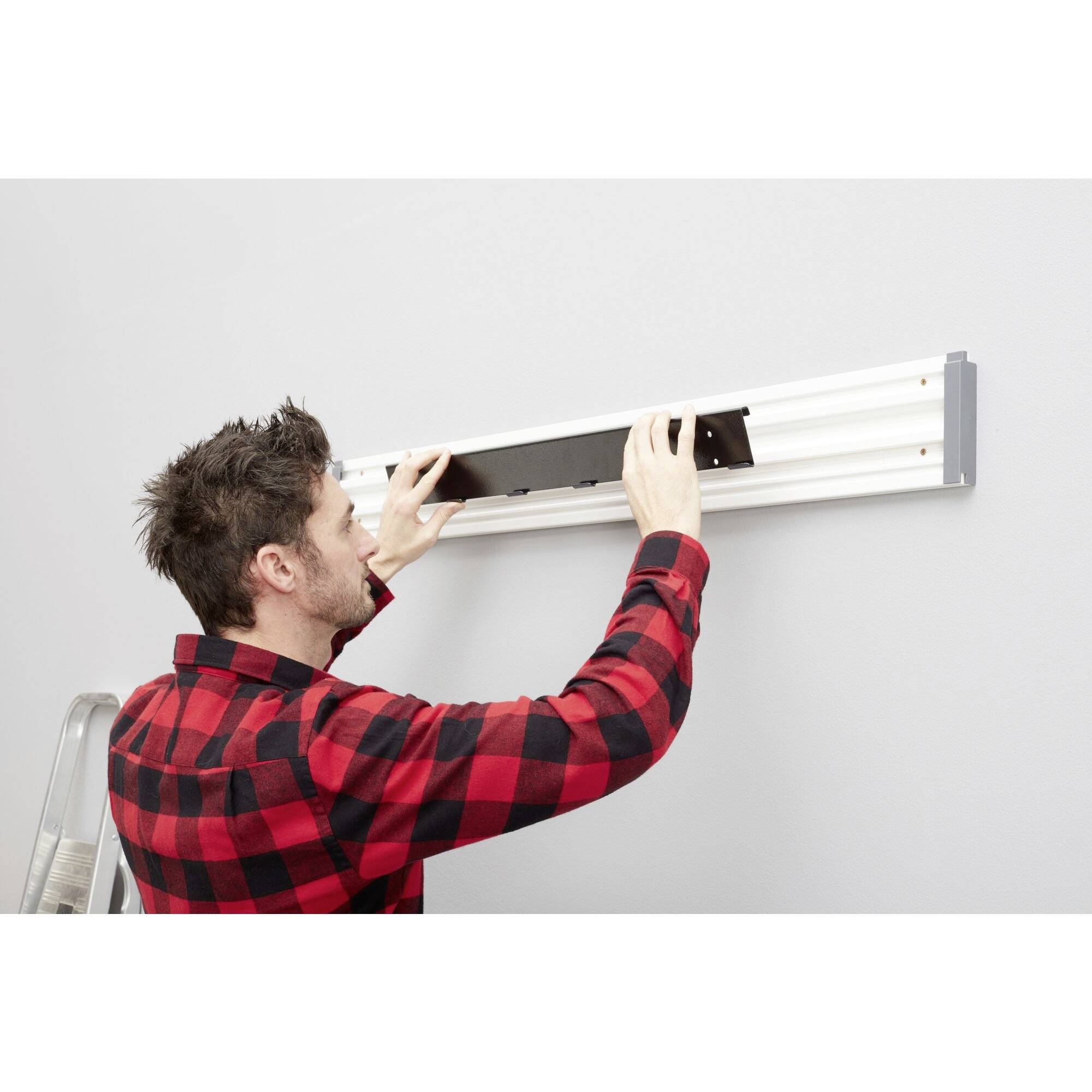 Un homme portant une chemise à carreaux monte une étagère sur un mur gris, debout sur une échelle.