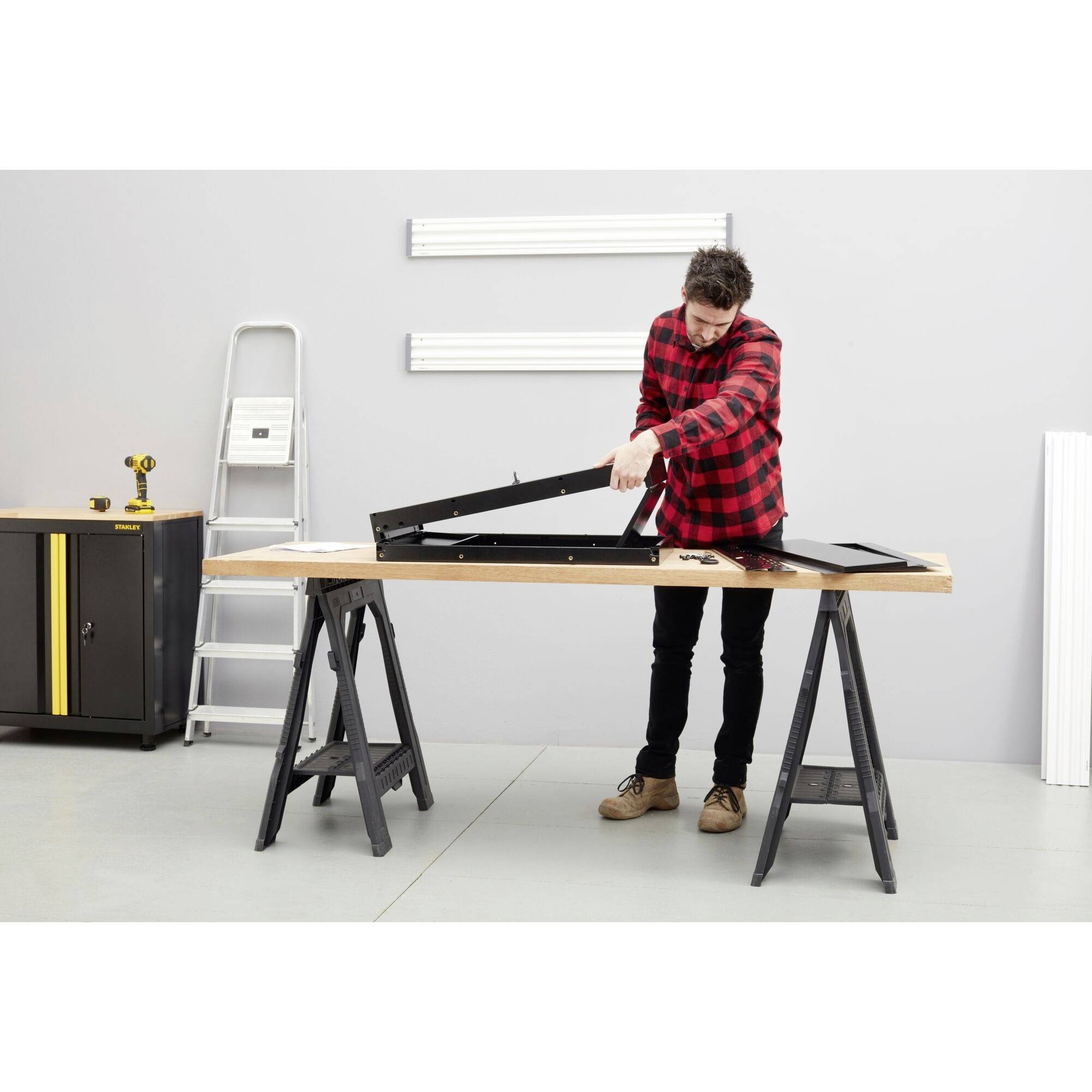 Une personne en vêtements de travail installe un châssis métallique noir sur une table d'atelier, avec des échelles et des outils en arrière-plan.