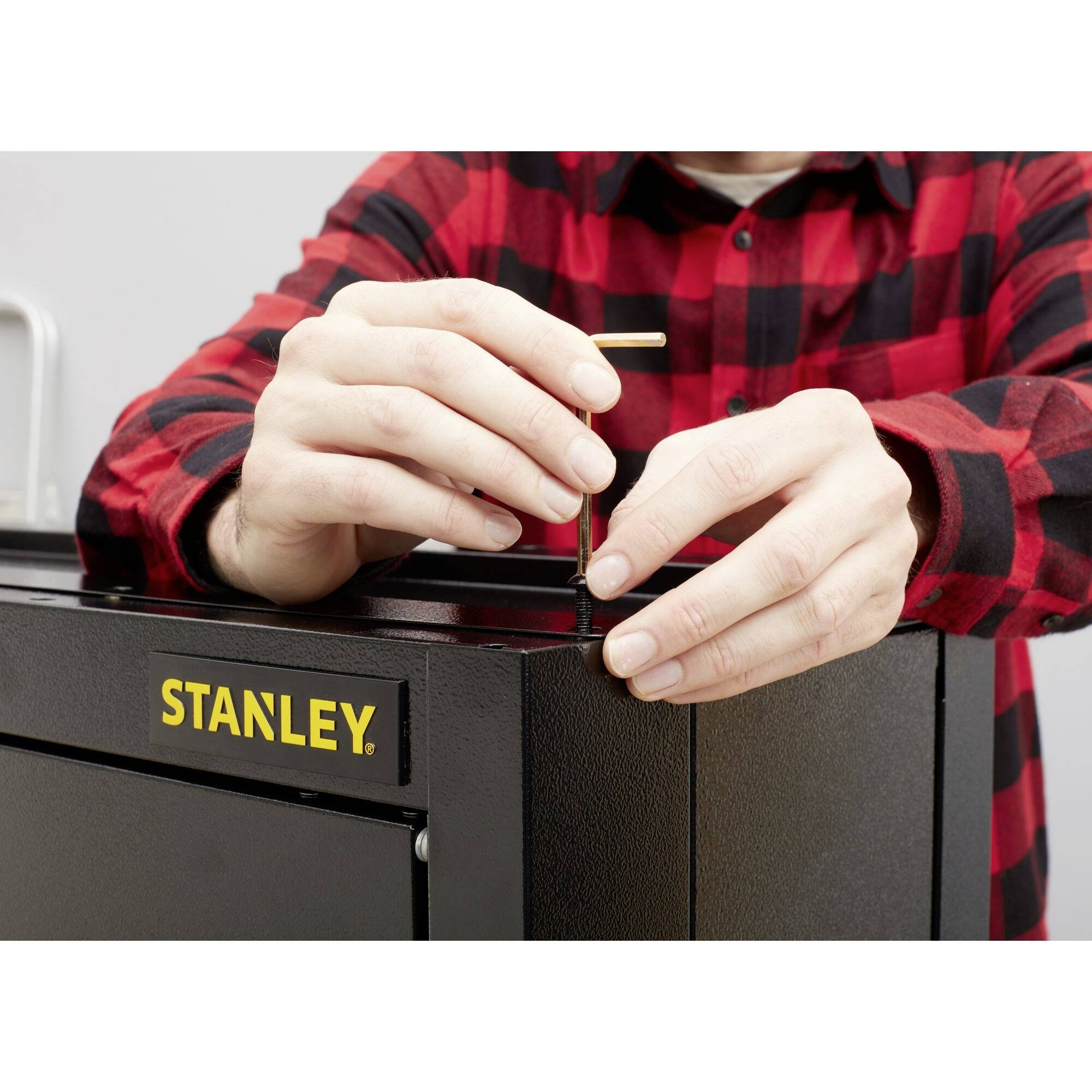 Une personne portant une chemise à carreaux rouge et noir installe un meuble à outils noir de Stanley. Elle utilise un petit outil métallique.