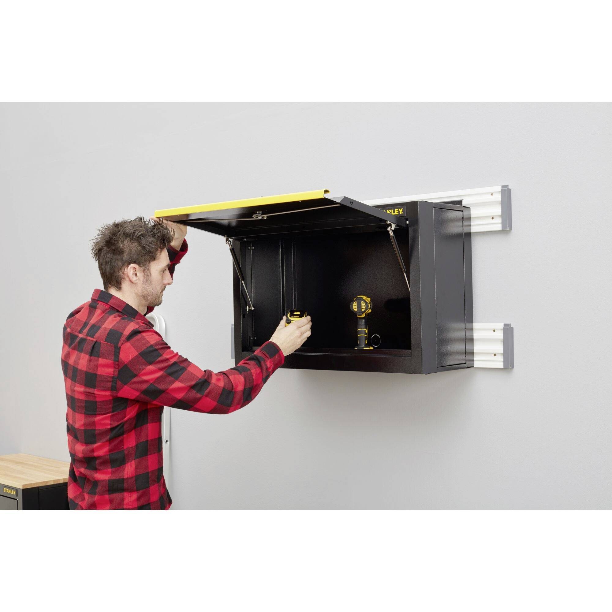 Un homme portant une chemise à carreaux rouges ouvre un placard noir et en sort un outil. L'armoire est fixée à un mur gris.