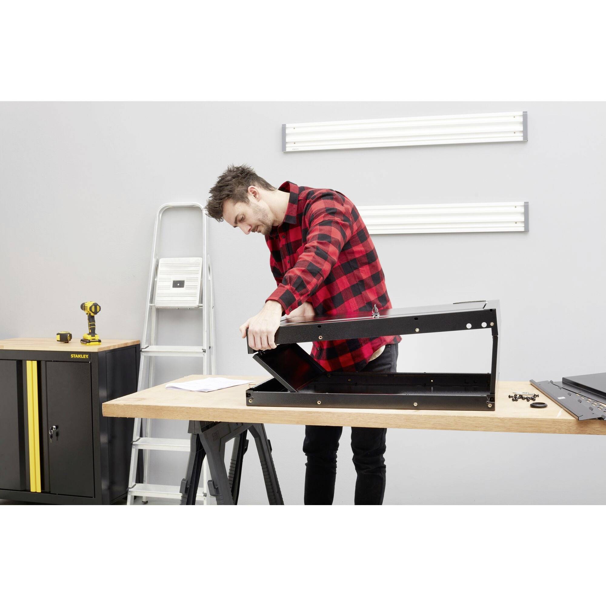 Un homme portant une chemise à carreaux rouge et noir monte un meuble sur une table en bois, avec une échelle et des outils en arrière-plan.