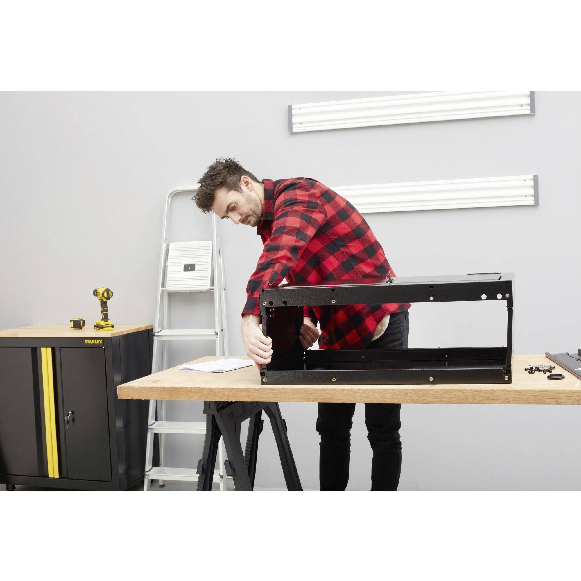 Une personne portant une chemise à carreaux rouge et noir monte un meuble noir sur un établi dans un environnement d'atelier.