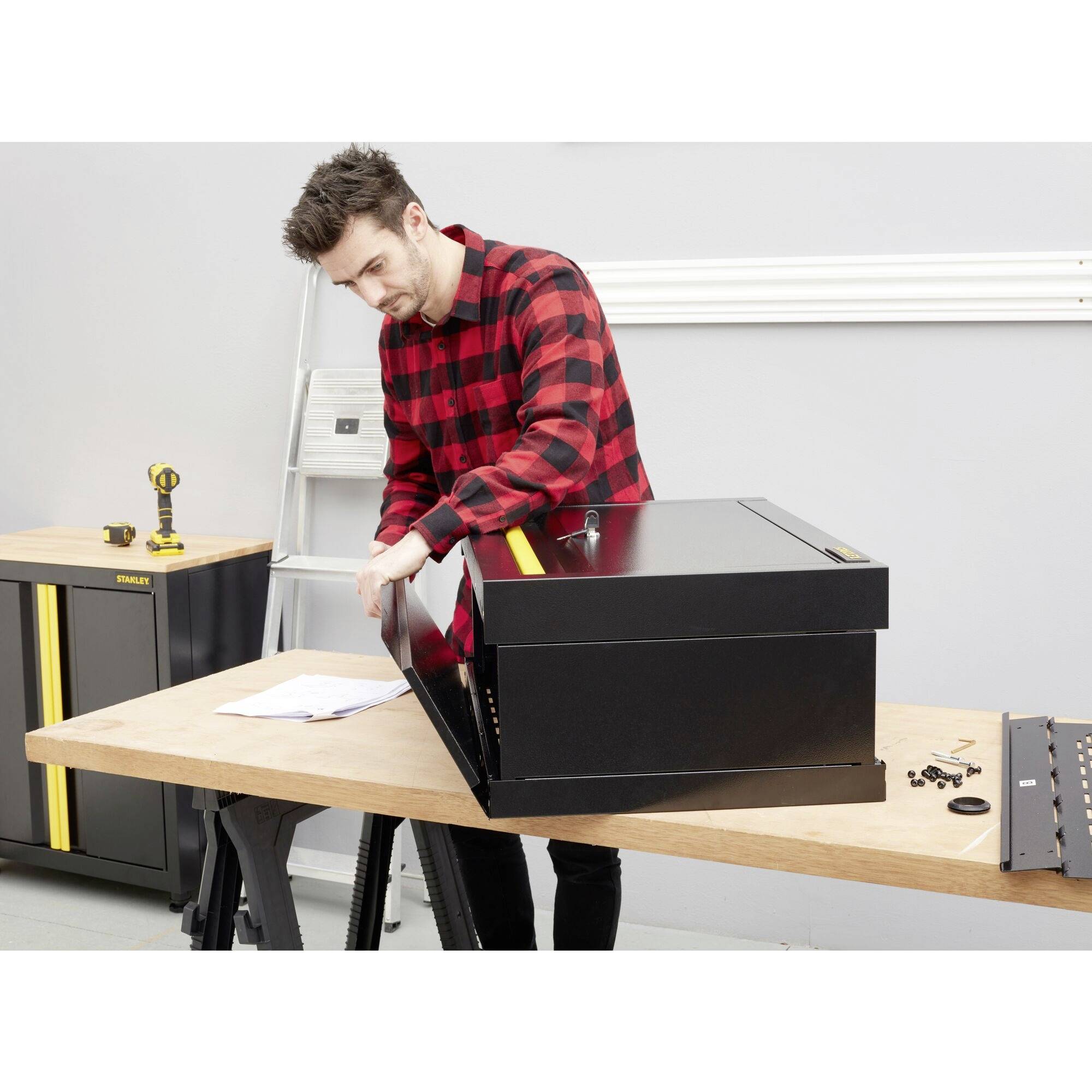 Un homme portant une chemise à carreaux assemble une boîte à outils noire dans un atelier. Des outils sont visibles en arrière-plan.