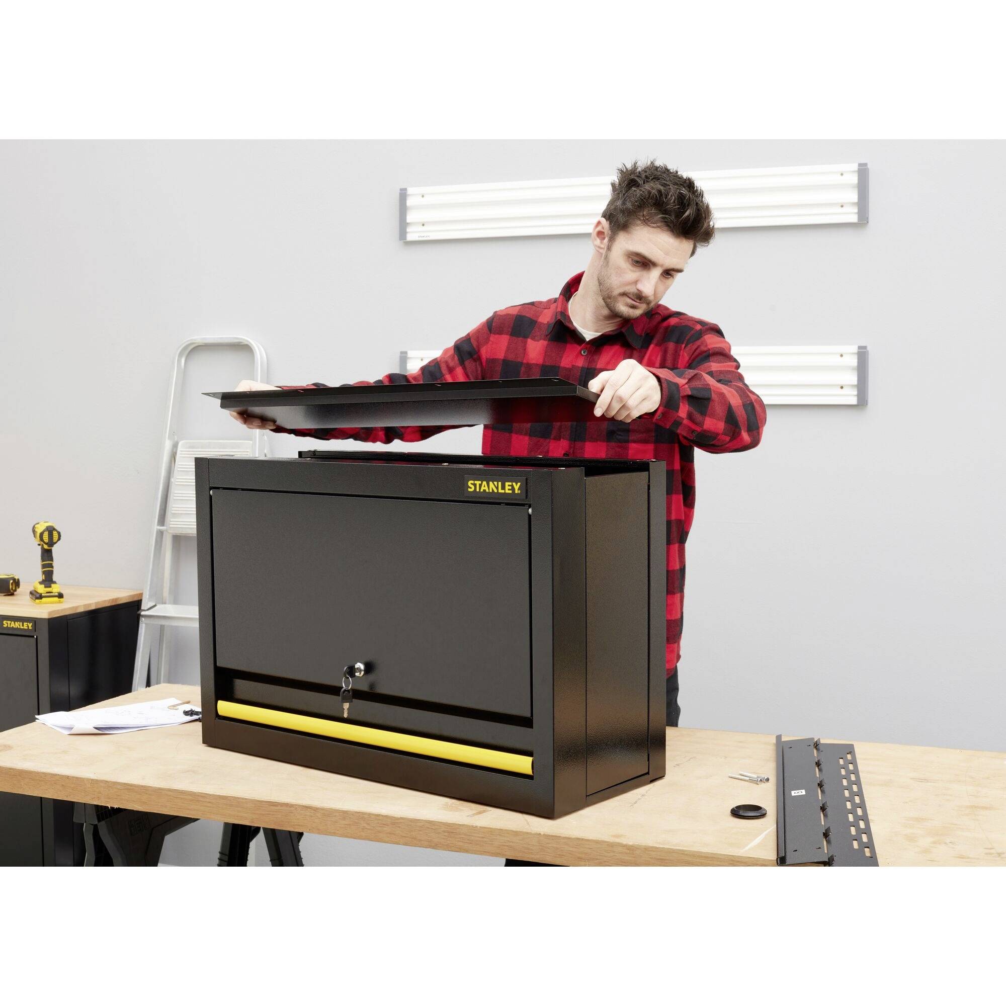 Un homme portant une chemise à carreaux rouge et noir installe une boîte à outils noire avec un détail jaune sur un établi.
