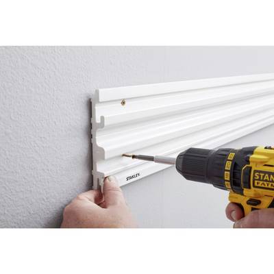 Une personne fixe une baguette blanche en plastique sur un mur gris à l'aide d'un tournevis sans fil jaune.