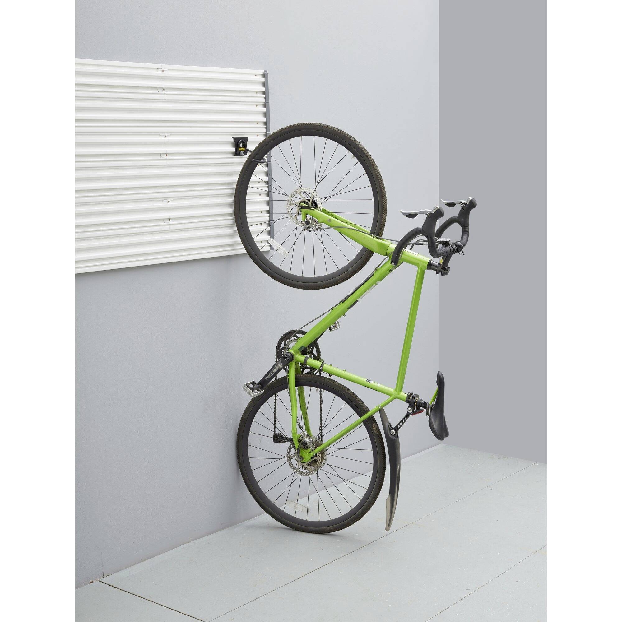 Un vélo vert est fixé verticalement contre un mur gris, sa roue avant suspendue en haut. Le sol de la pièce est clair.