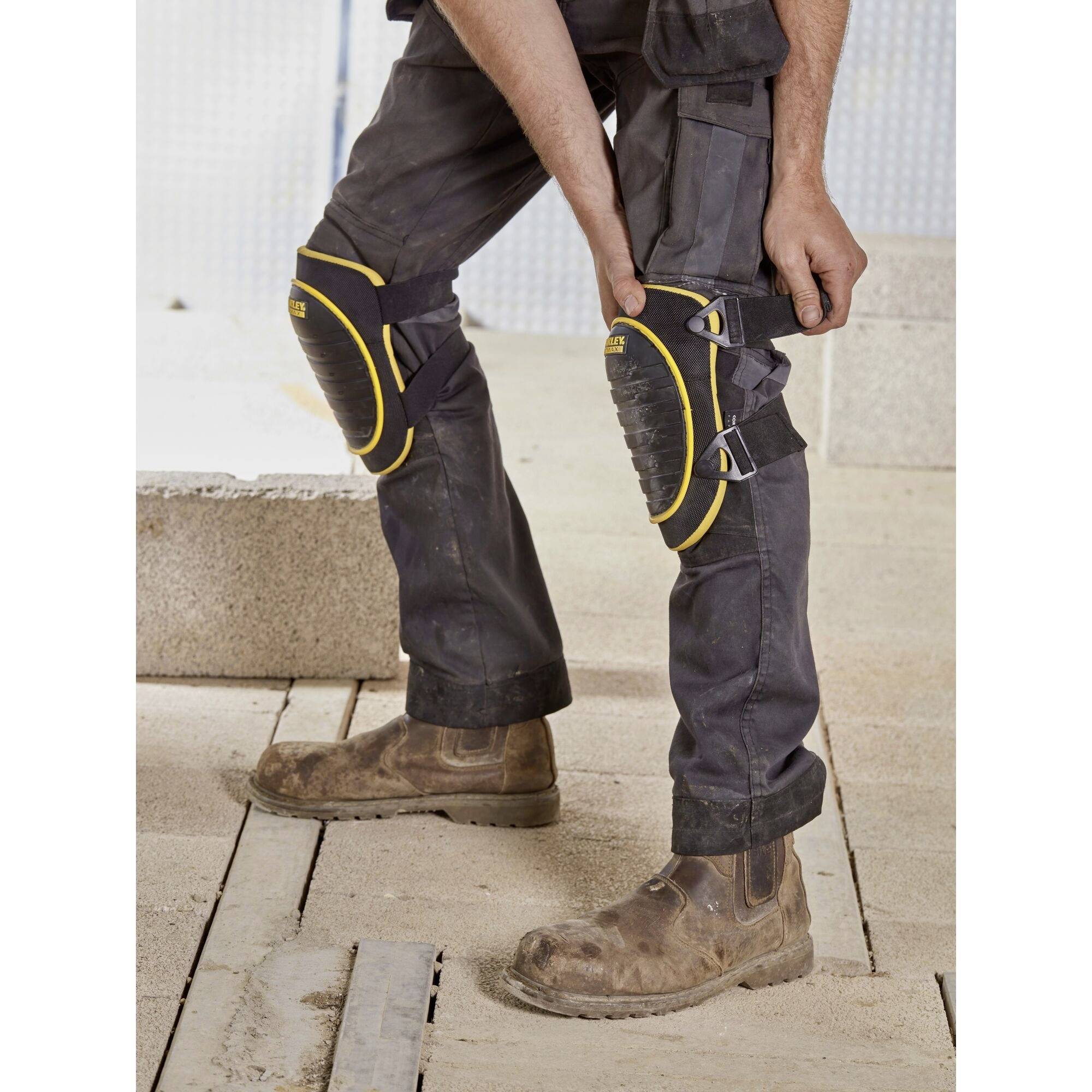Une personne porte un pantalon de travail noir, des bottes brunes et des protège-genoux jaunes et noirs, debout sur un sol en béton dans un environnement de chantier.
