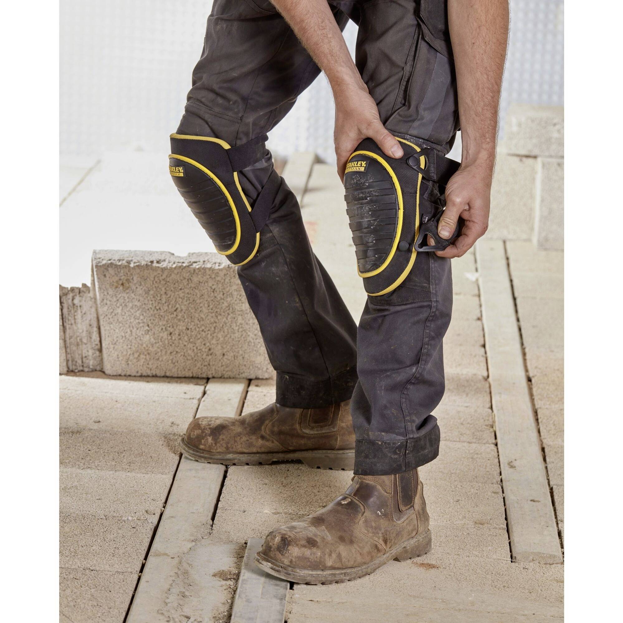 Un ouvrier portant des genouillères et un pantalon de travail se tient sur un chantier à côté de blocs de béton empilés.