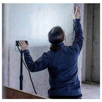 Une personne vêtue d'une veste sombre mesure ou marque un mur en béton avec un appareil portatif, se tenant à l'intérieur près de surfaces de construction inachevées.