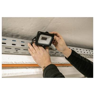 Des mains installant un projecteur LED noir sur un support de plafond ou de mur au-dessus d'un chemin de câbles métallique.