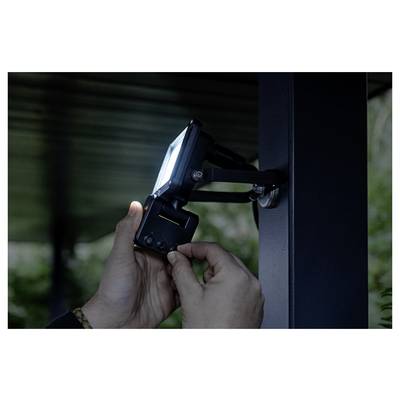 Les mains ajustent un projecteur de sécurité extérieur monté sur un poteau métallique, avec la lampe orientée vers la gauche en plein jour.