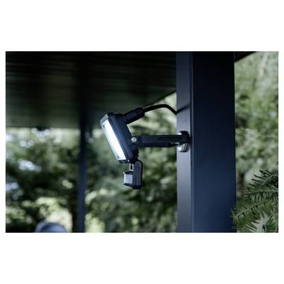 Une lumière de sécurité LED montée sur un poteau sombre sous un toit, orientée vers l'extérieur avec des arbres flous en arrière-plan.