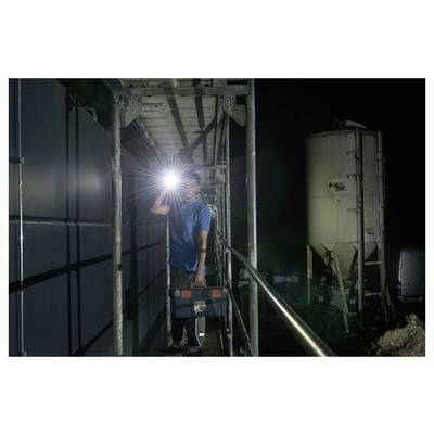 Ouvrier debout sur un échafaudage la nuit, tenant une lampe de poche brillante dans une zone industrielle à côté d'un grand réservoir et de véhicules garés.