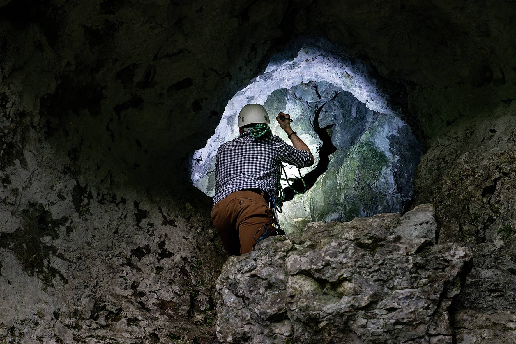 Une personne portant un casque et un équipement d'escalade explore une grotte, regardant à travers une petite ouverture dans la roche, entourée de pierre sombre.