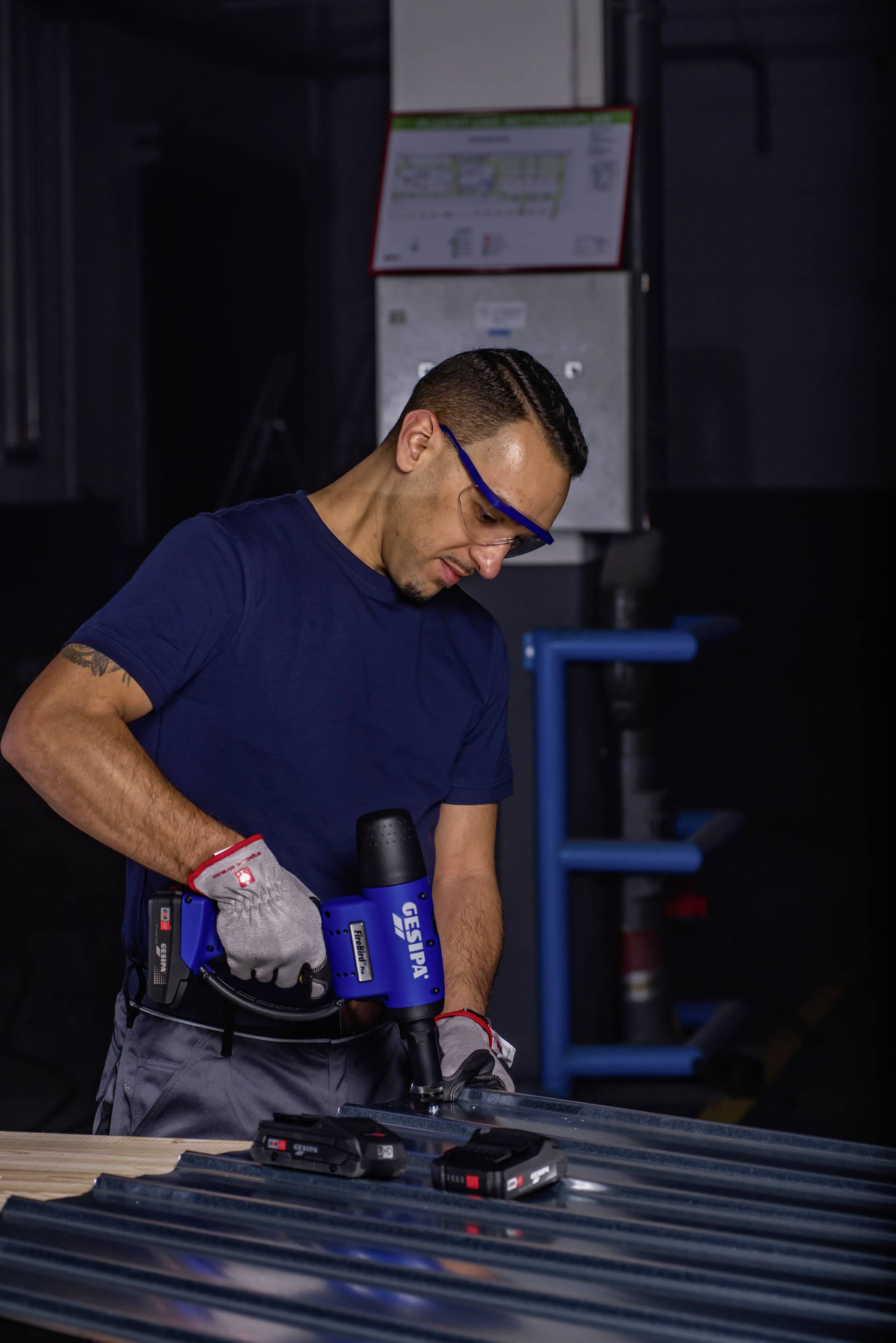 Une personne portant des lunettes de protection et des gants utilise un outil électrique pour travailler une grande tôle dans un environnement industriel.
