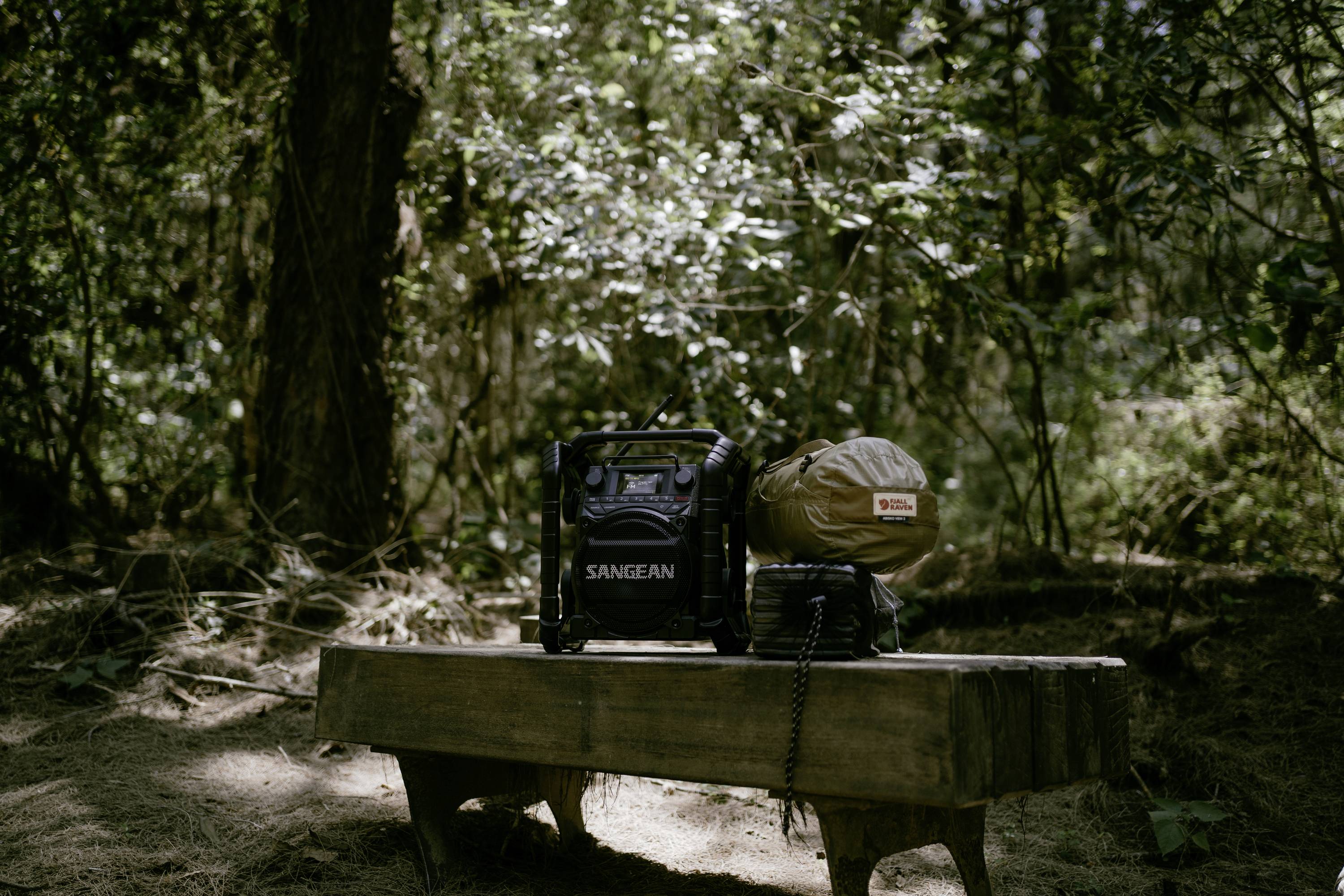 Une radio et un sac sont posés sur un banc en bois dans la forêt. La lumière du soleil traverse les arbres.