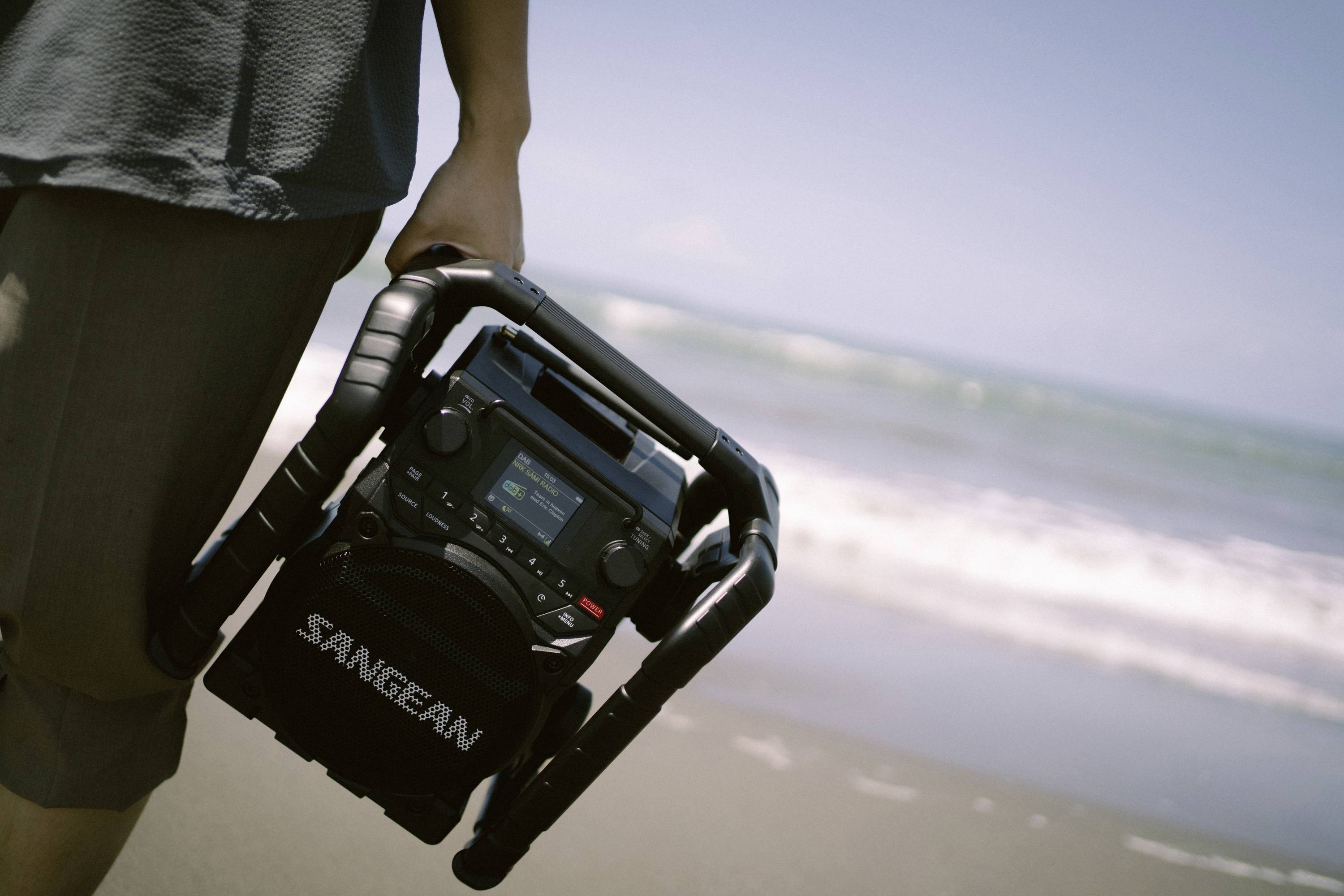 Une personne tient une radio portable de marque Sangean sur la plage. En arrière-plan, on peut voir la mer et les vagues.