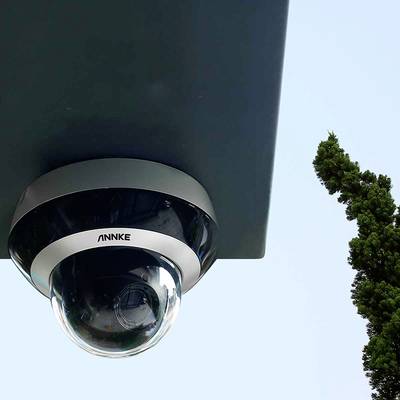 Caméra de surveillance montée au plafond, avec un arbre vert à l'arrière-plan sous un ciel bleu.