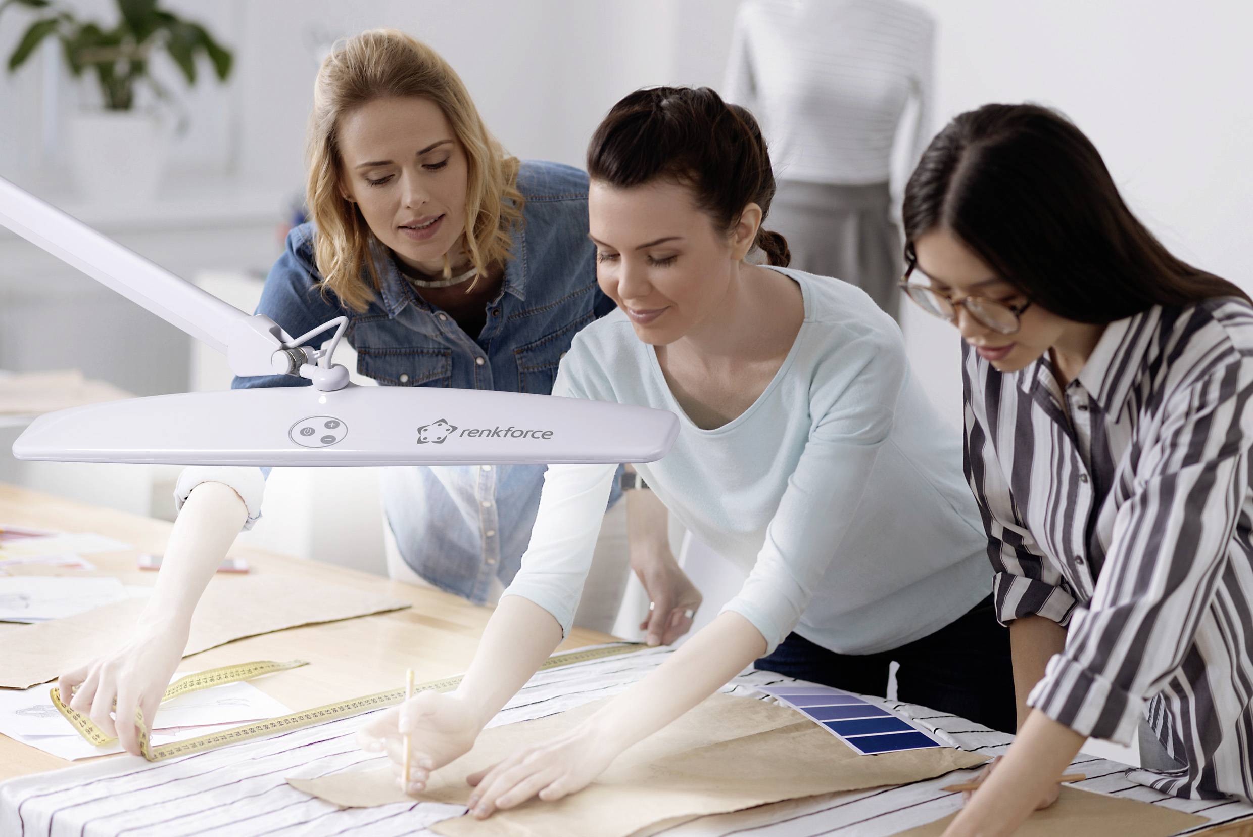 Trois femmes travaillent sur des échantillons de tissus et des patrons autour d'une table. Une lampe de bureau éclaire leur espace de travail.