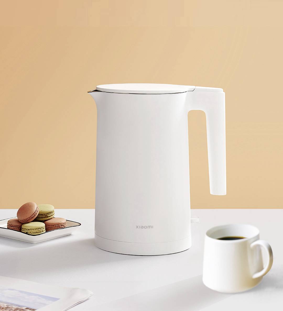 Une bouilloire blanche est posée sur une table, à côté d'une pile de macarons et une tasse de café. L'arrière-plan est dans un ton orange chaleureux.
