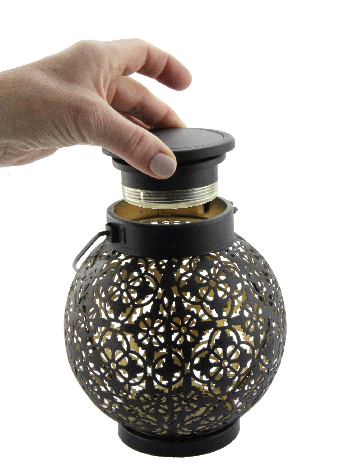 Une main retire le couvercle d'une lanterne décorative ronde au motif ajouré. La lanterne est noire et possède un insert en verre.