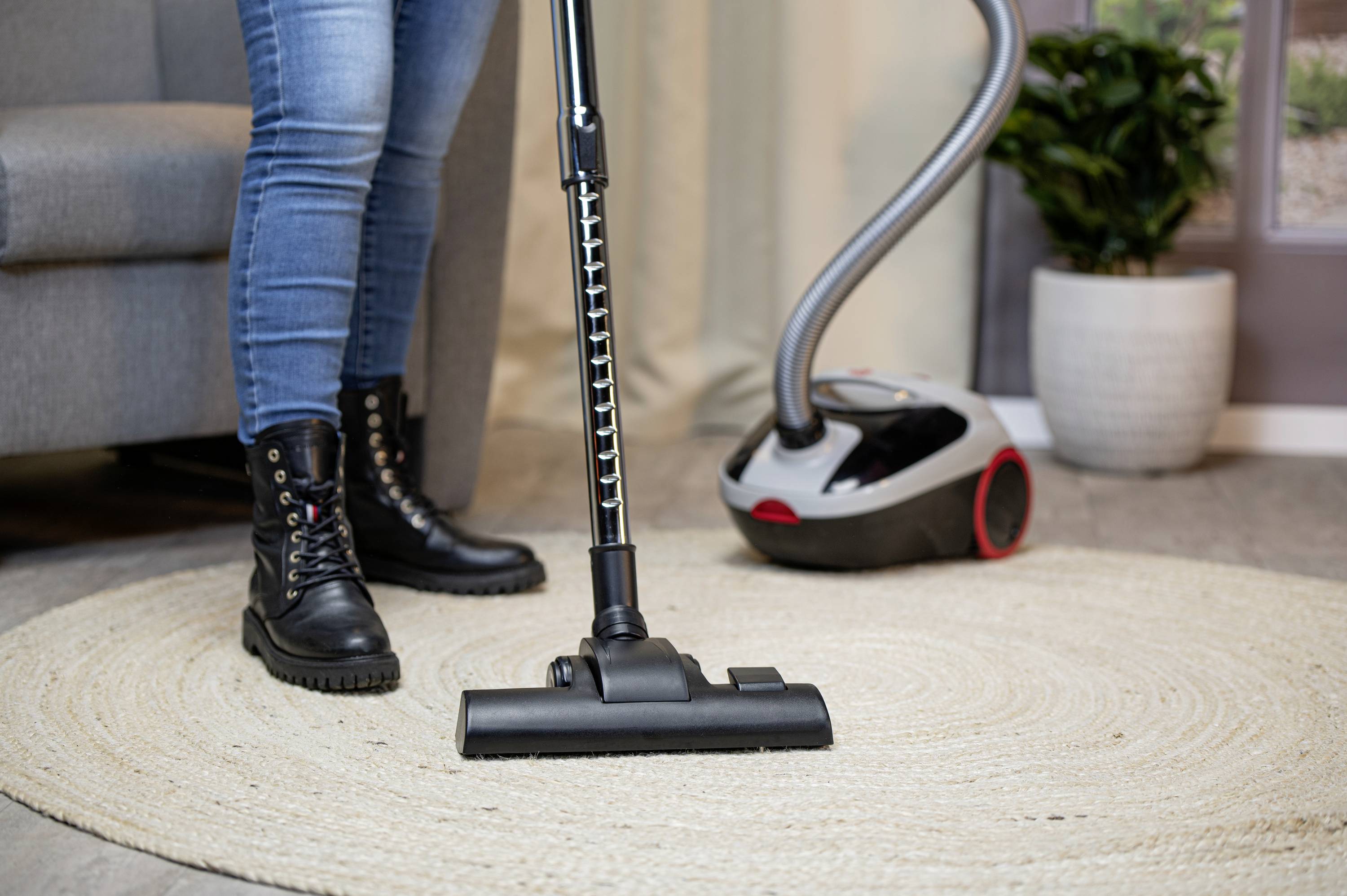 Une personne en jeans et bottes aspire un tapis rond à l'aide d'un aspirateur. Un canapé et une plante sont visibles en arrière-plan.
