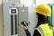 Une personne portant un casque de protection et un gilet de sécurité vérifie un boîtier électrique dans un bâtiment à l'aide d'un appareil de test électrique.