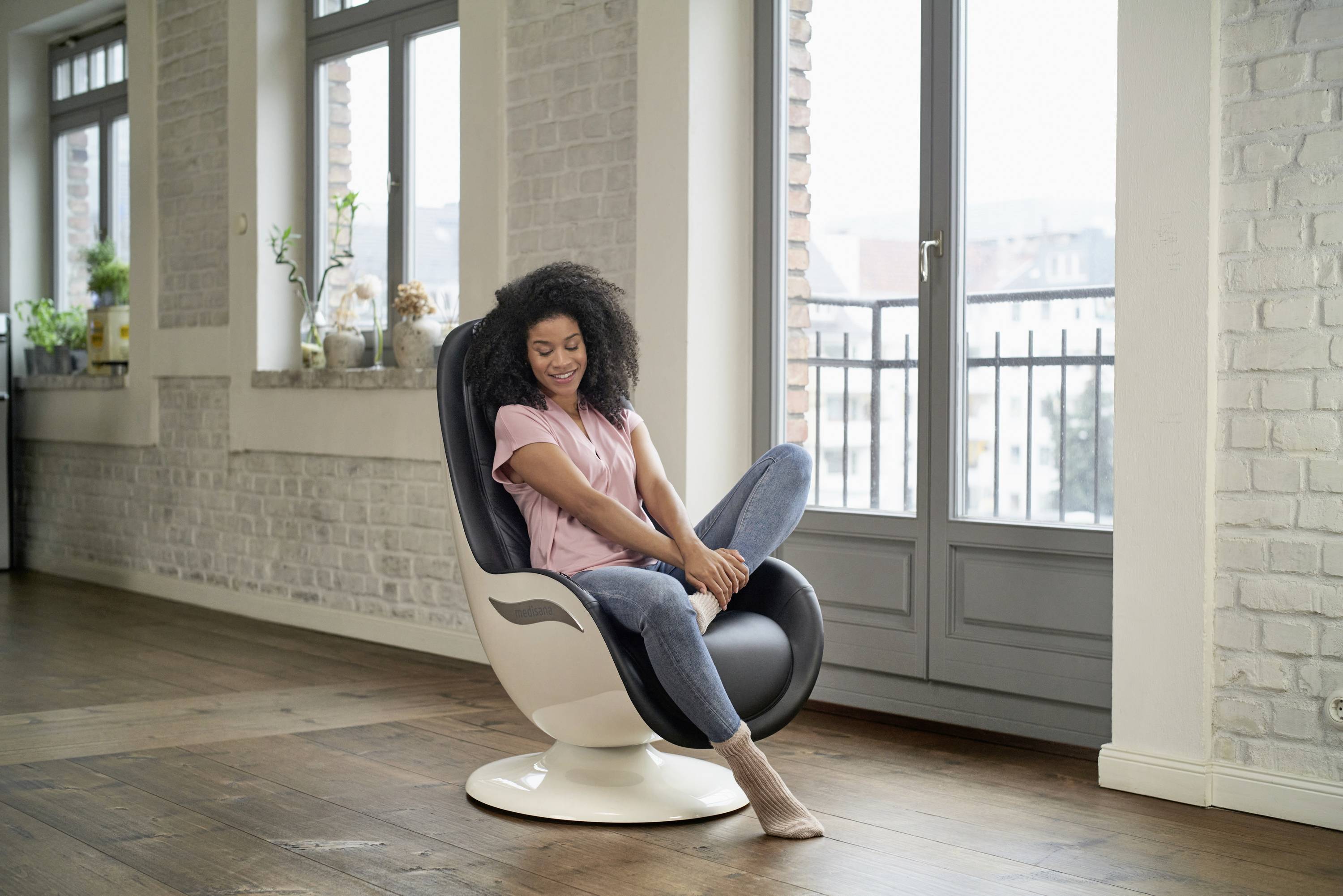 Une femme est assise de manière détendue sur un fauteuil moderne dans une pièce baignée de lumière, avec de grandes fenêtres et un sol en bois.