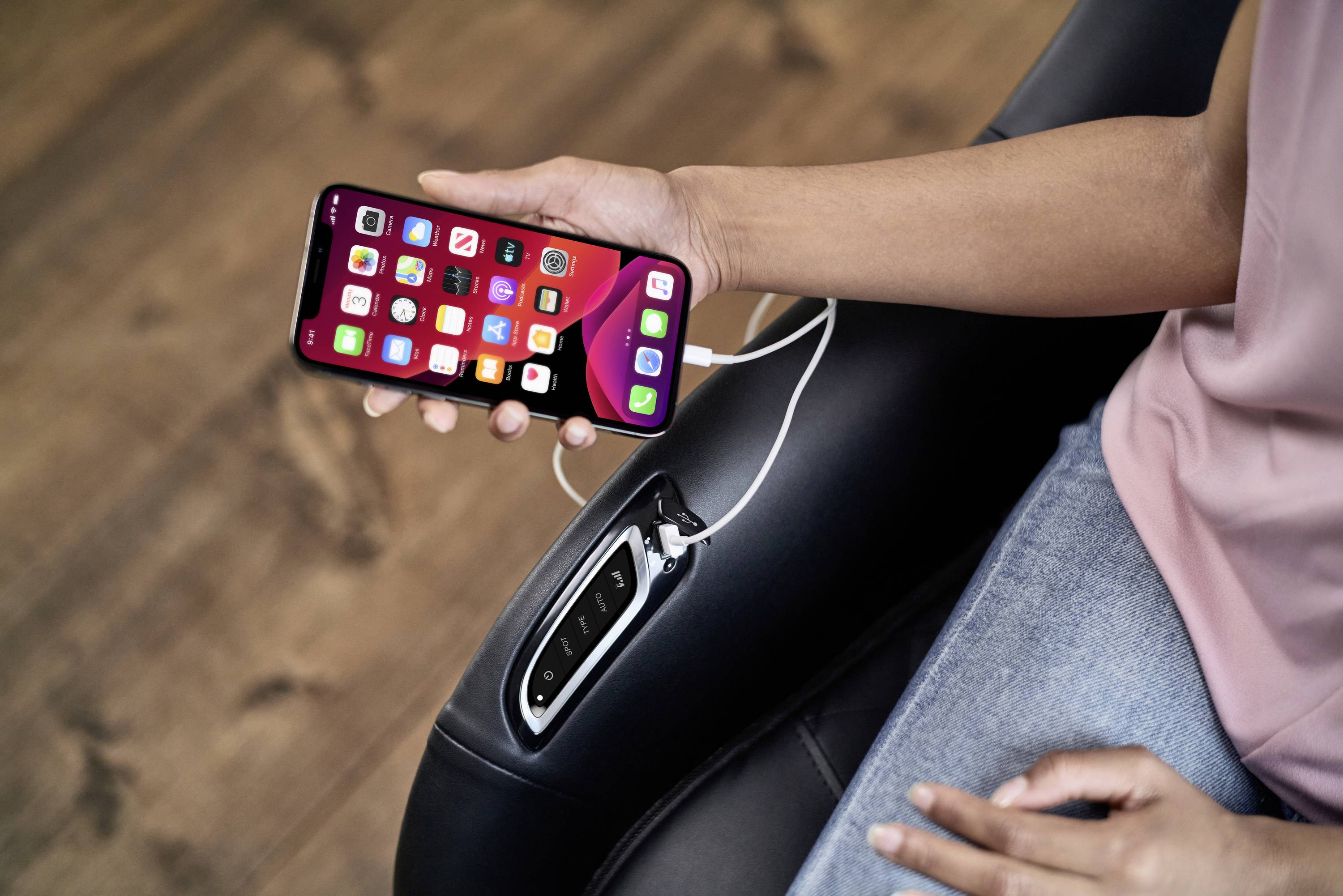 Une personne tient un smartphone dans sa main, qui est posé avec son câble de charge sur un fauteuil. L'écran affiche des applications.