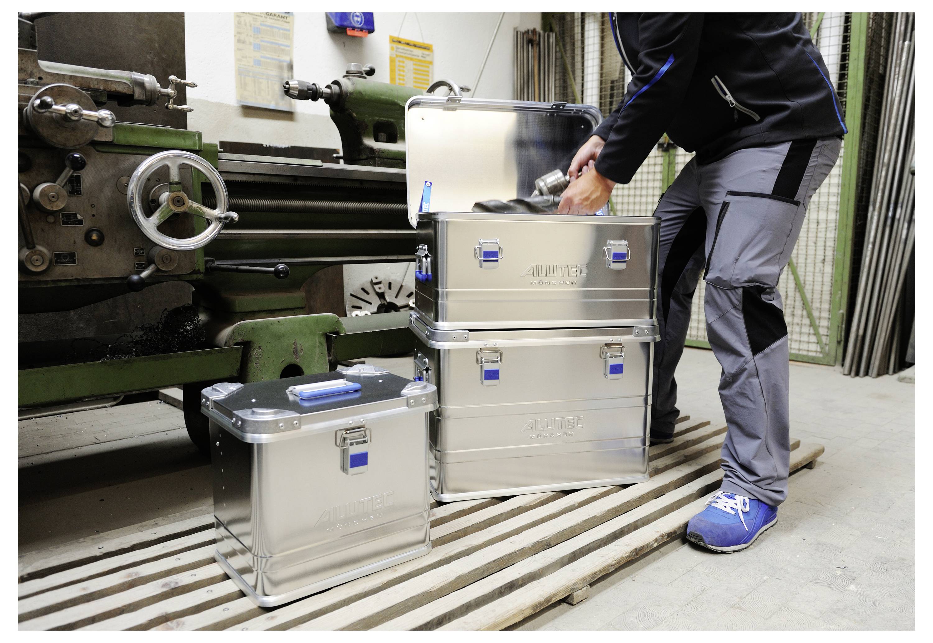 'Une personne empilant des boîtes de rangement en métal dans un atelier, à proximité d'une grande machine industrielle, organisant les outils et l'équipement.'