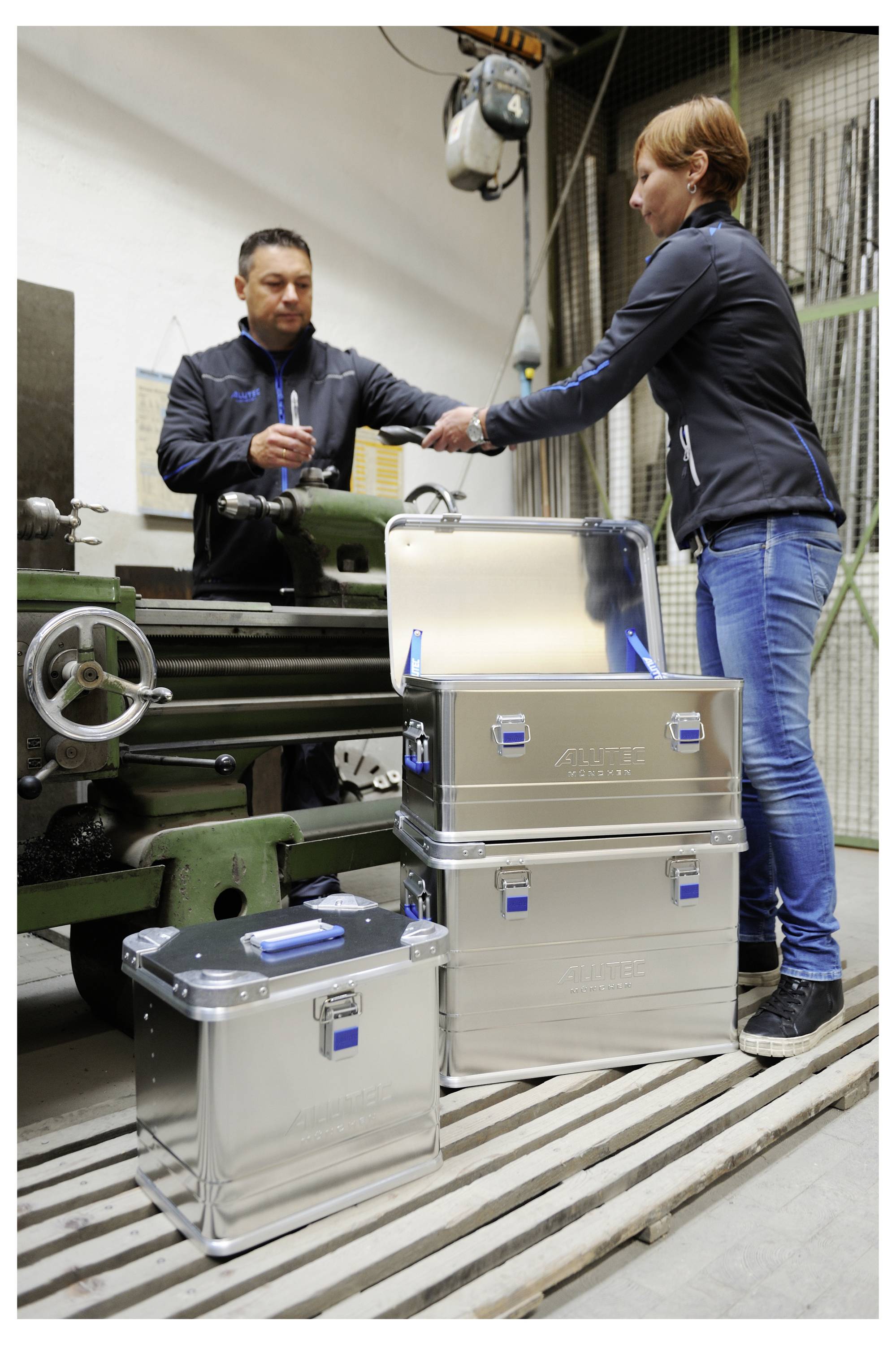 Un homme et une femme dans un atelier se tiennent près d'une machine métallique, regardant des documents. Trois boîtes de stockage argentées sont empilées à proximité.
