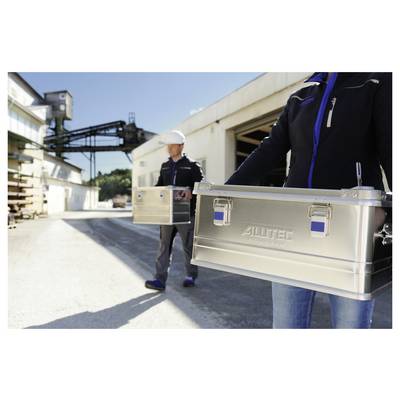 Deux personnes transportant des coffres à outils en métal portant le label 'ALUTEC' à l'extérieur sur un site industriel, avec des bâtiments et des machines en arrière-plan.