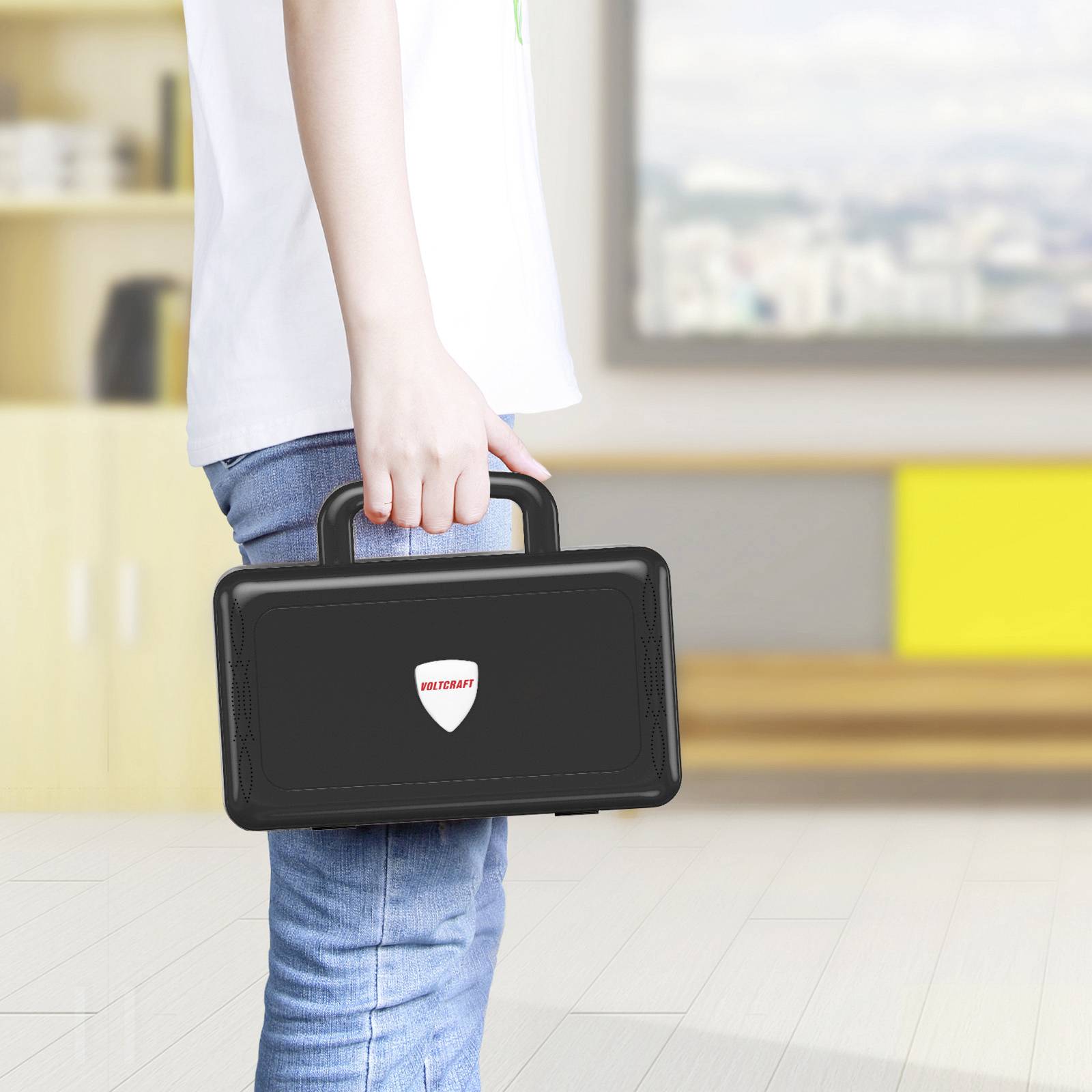 Une personne porte une valise noire avec un autocollant en forme de cœur dans une pièce claire. Vue de dos, avec l'accent mis sur la valise.