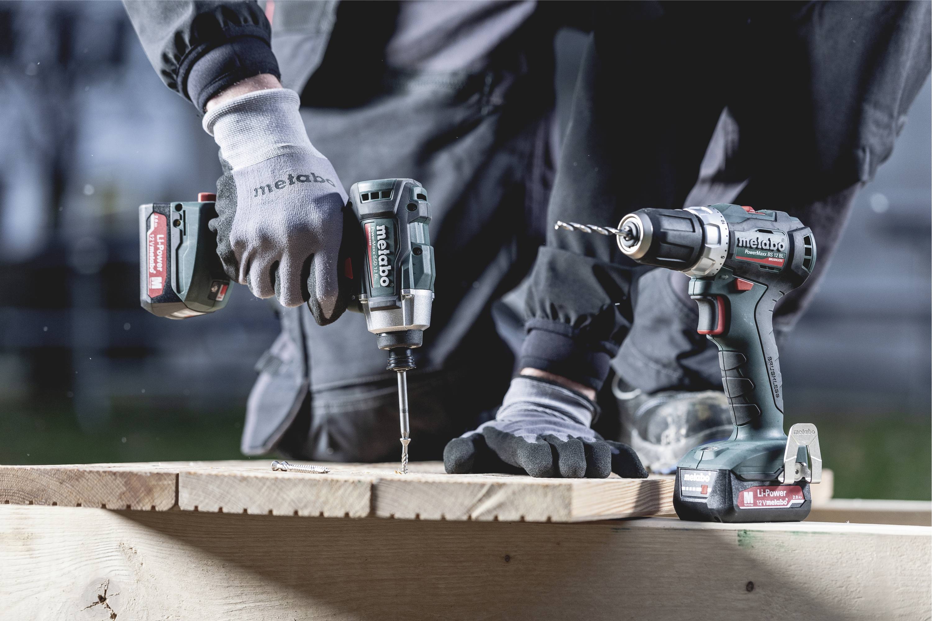 Une personne en vêtements de travail utilise une visseuse sans fil pour visser une vis dans une planche en bois. Une deuxième visseuse sans fil est posée à côté.