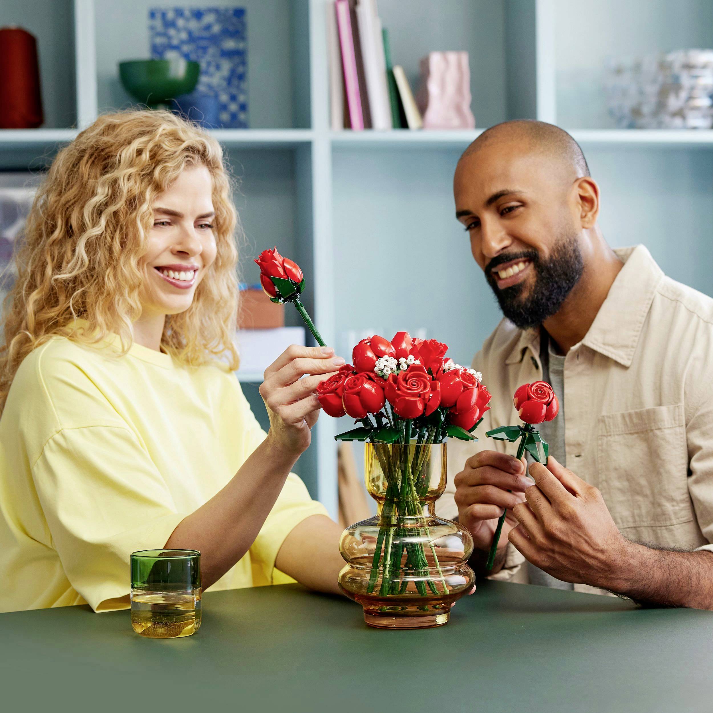 Une femme souriante et un homme sont assis à table, arrangeant des roses rouges dans un vase. À l'arrière-plan, on aperçoit une étagère décorée.