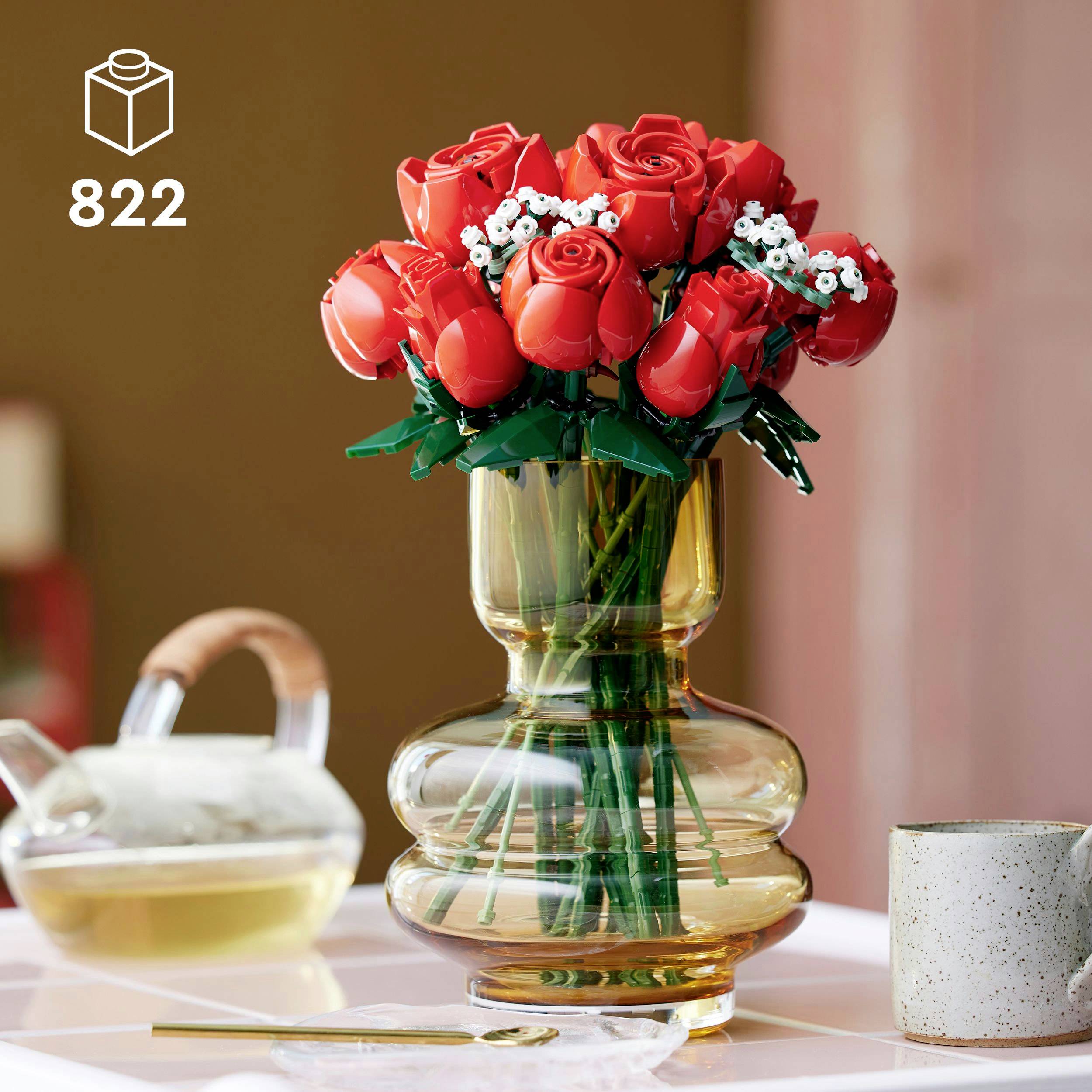 Un vase en verre contenant des roses en LEGO sur une table, à côté d'une théière de thé vert et d'une fourchette dorée.
