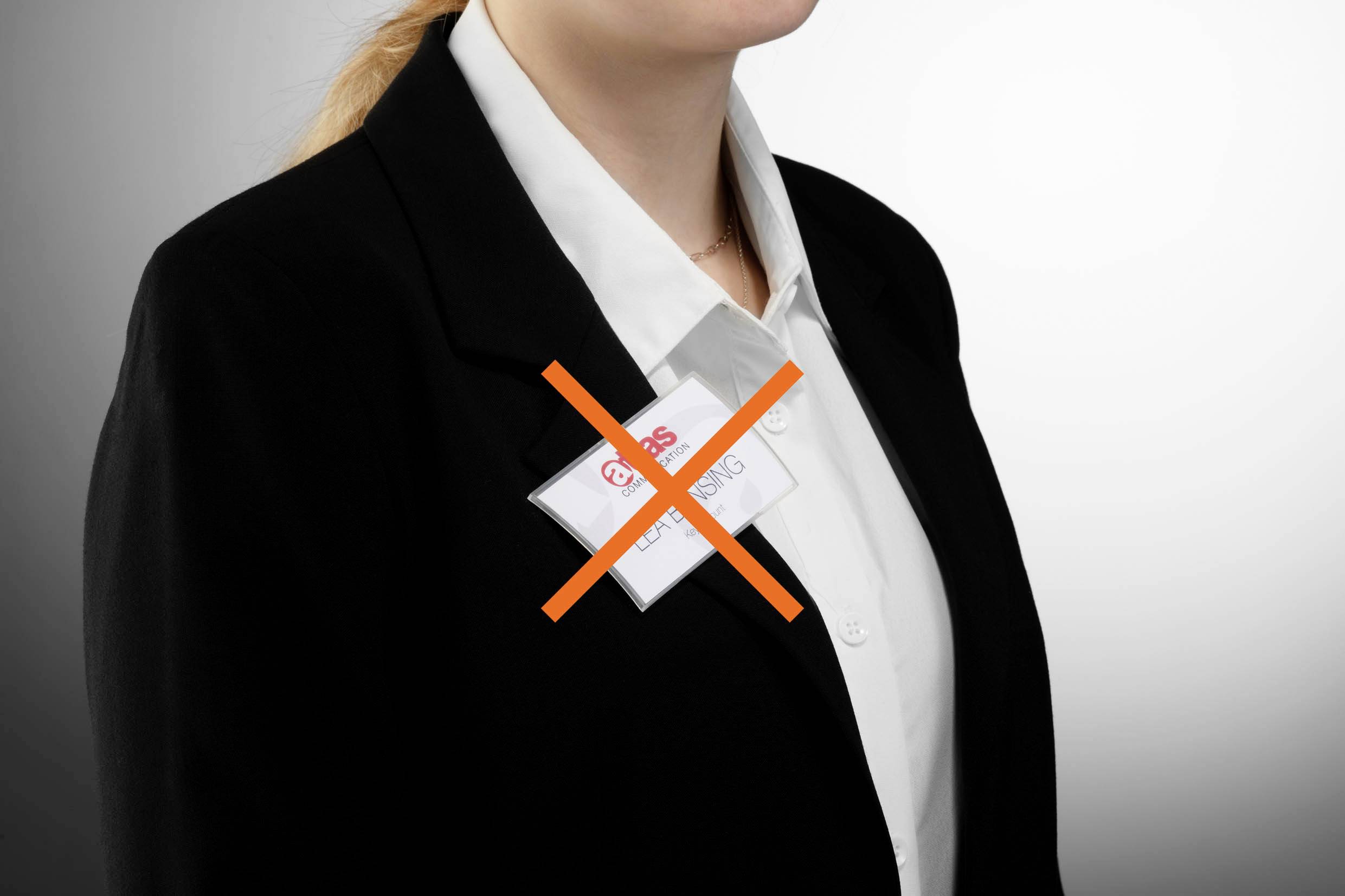 Une personne dans un costume noir avec un badge nominatif barré d'un X rouge, sur un fond gris.