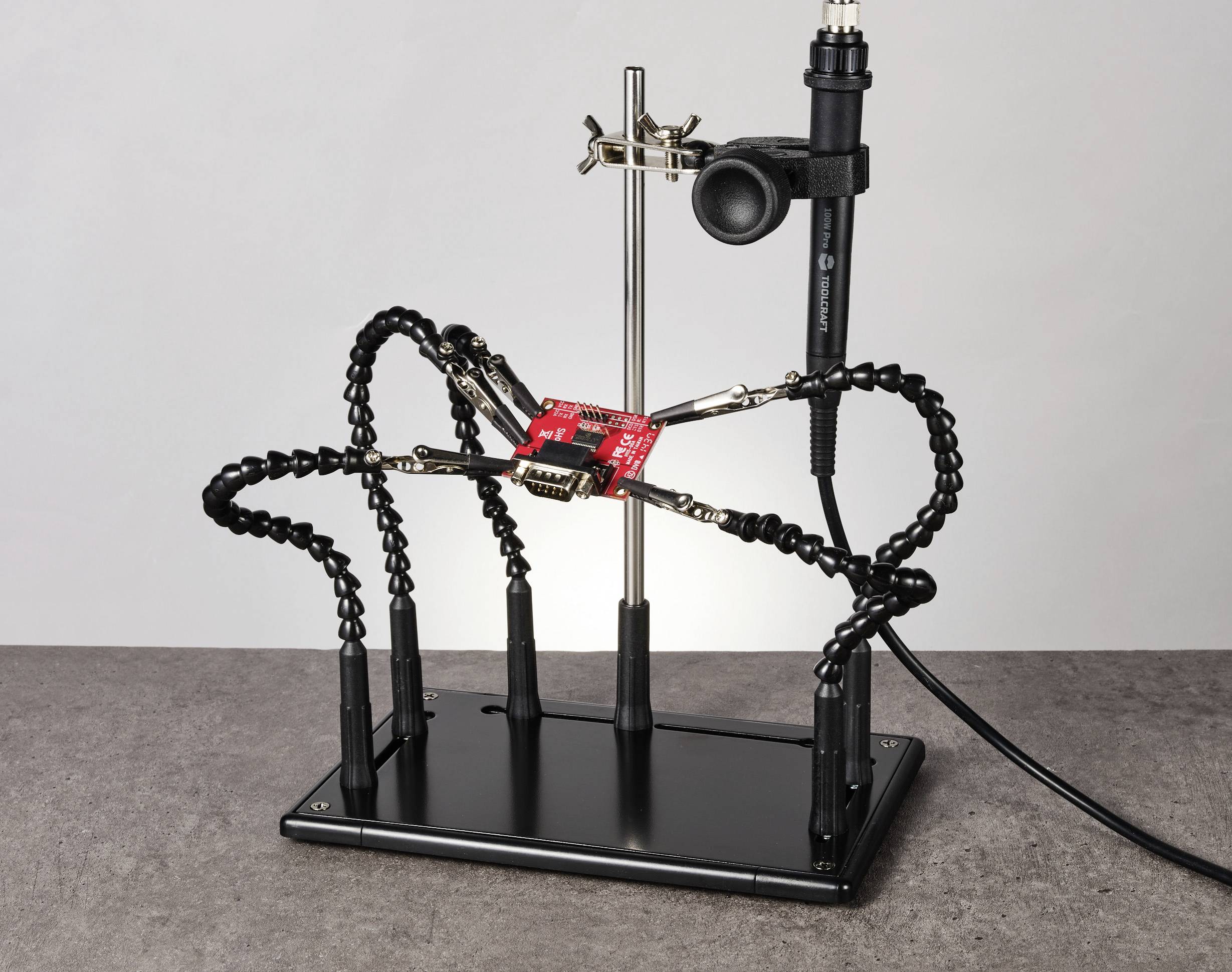 Un outil d'aide au soudage doté de bras flexibles maintient un circuit électronique rouge. Un fer à souder est fixé dans le support.