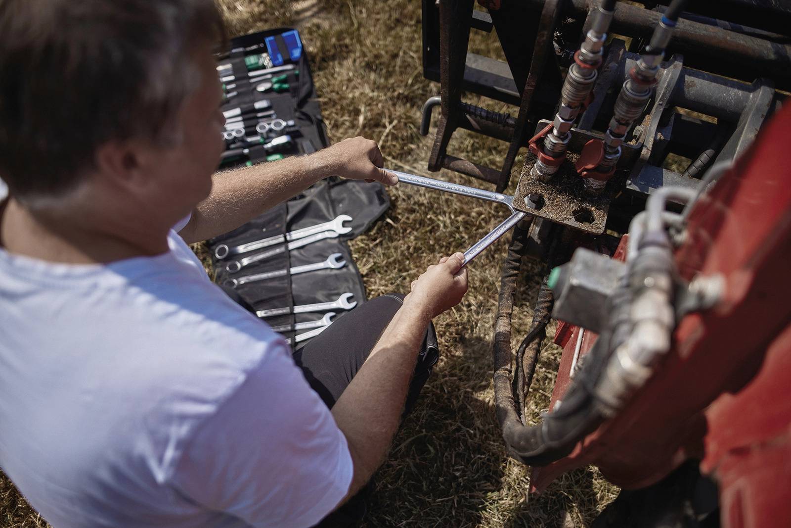 Un homme répare une machine avec des clés à molette dans un champ. Différents outils sont éparpillés autour de lui.