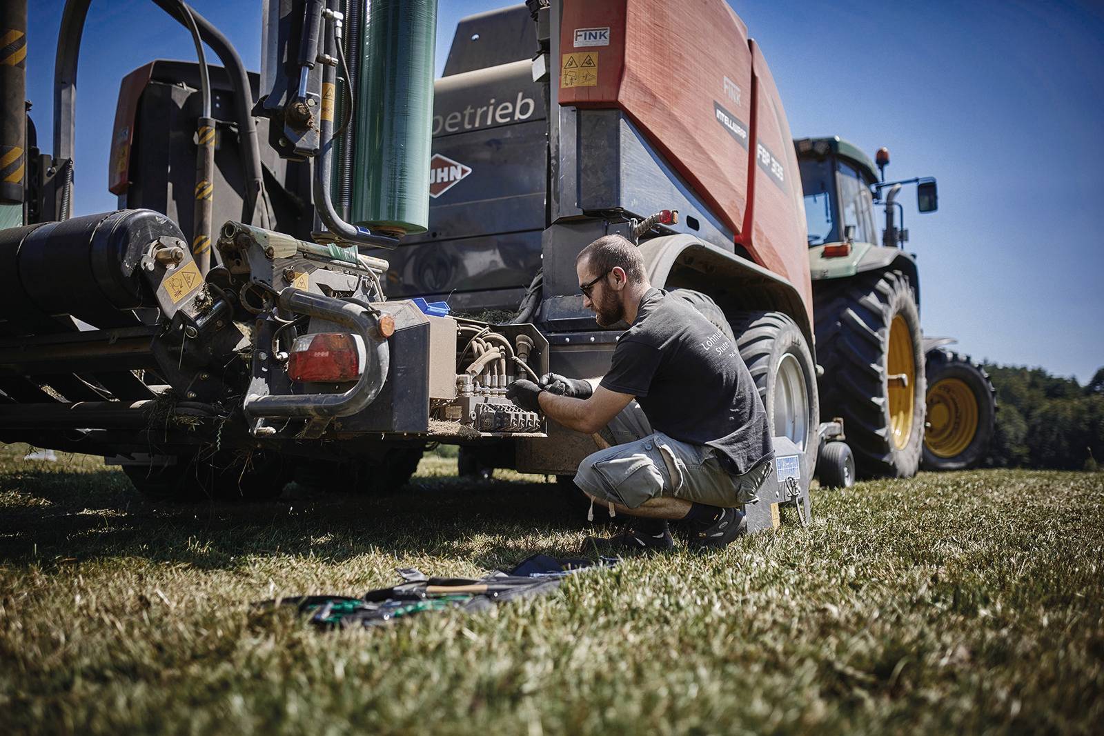 Un homme répare une grande machine agricole dans un champ en plein air. Le ciel est clair et bleu.