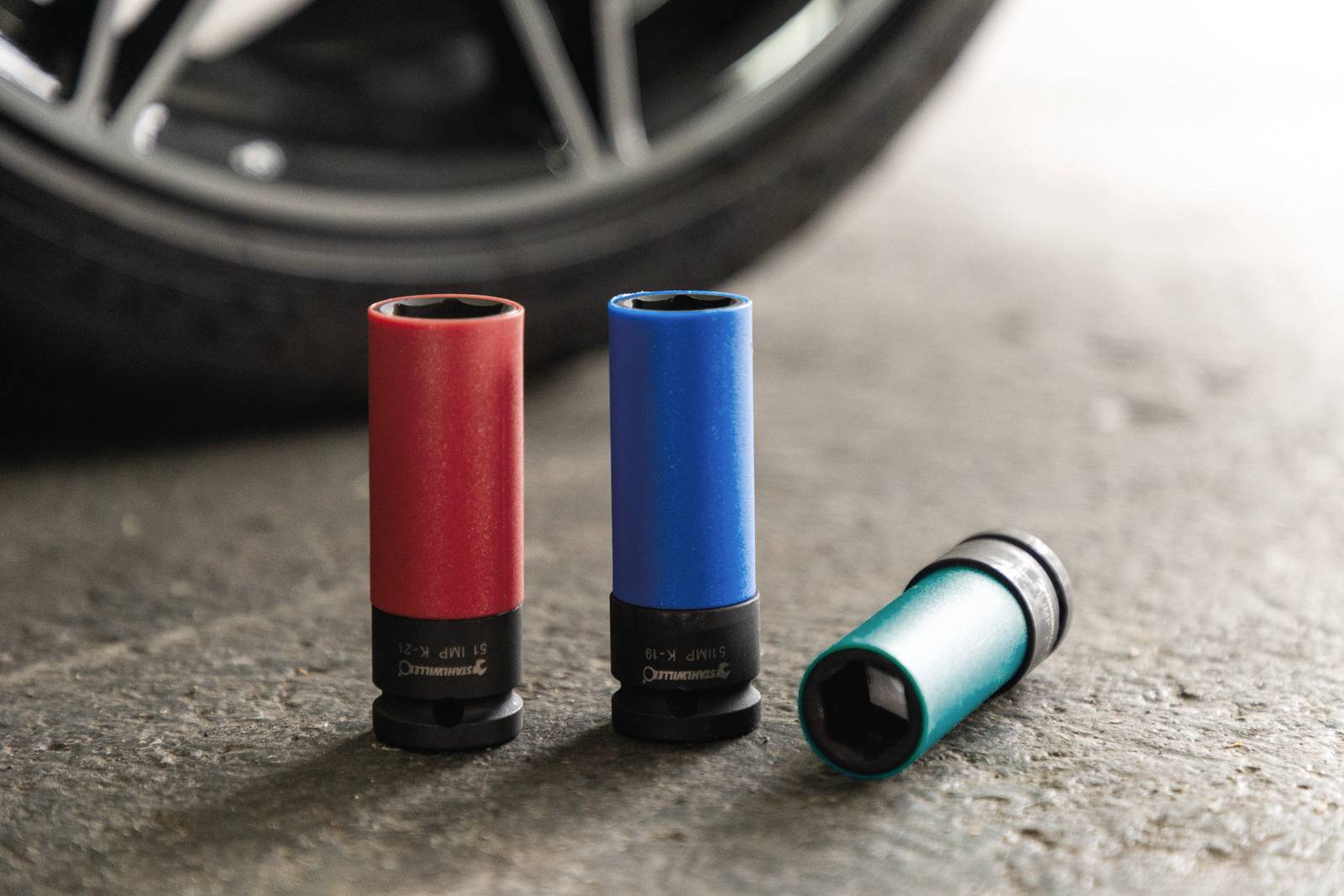 Trois douilles de clés colorées (rouge, bleu, vert) sont posées sur un sol en béton, près d'un pneu de voiture.