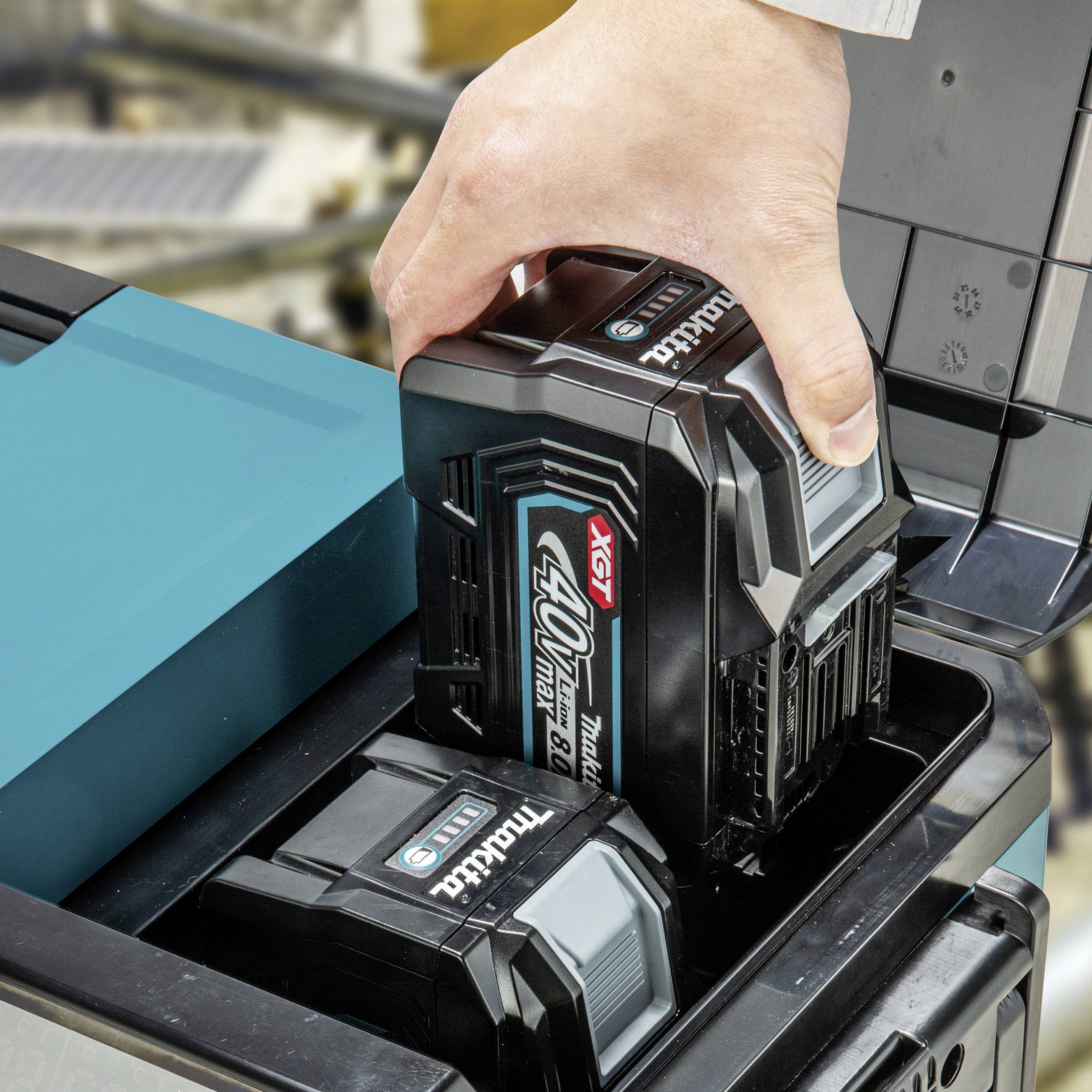 Une main insère une batterie de 40V dans une station de charge. Des outils sont visibles en arrière-plan, flous.