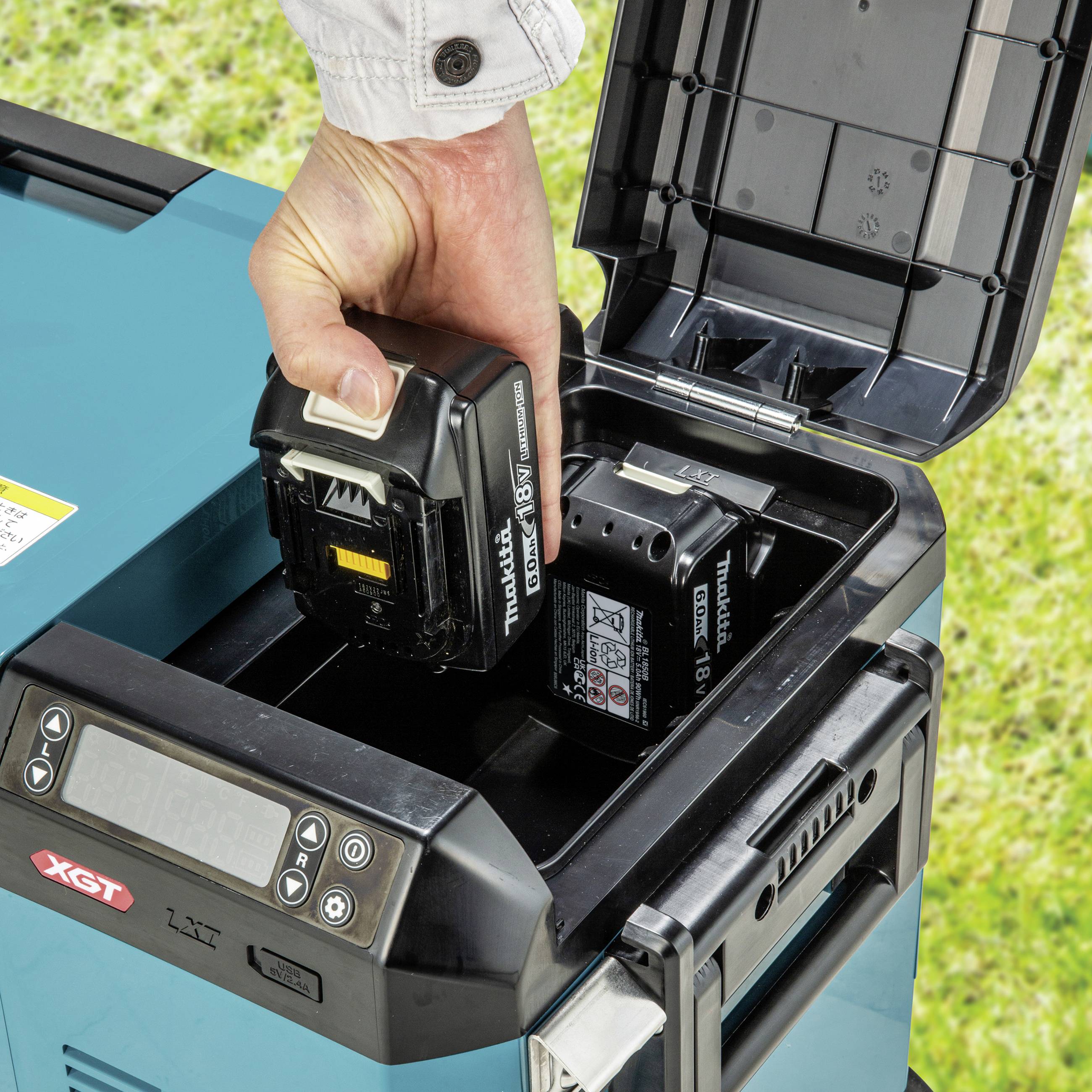 Une personne change une batterie dans un appareil électrique à l'extérieur. La batterie affiche la marque et la capacité. De l'herbe est visible en arrière-plan.