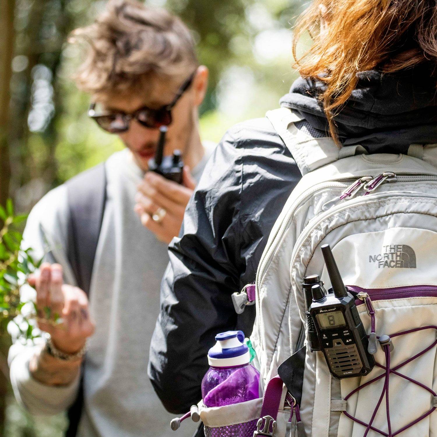 Deux personnes dehors ; l'une porte un sac à dos avec une radio attachée. L'environnement semble naturel et ensoleillé.